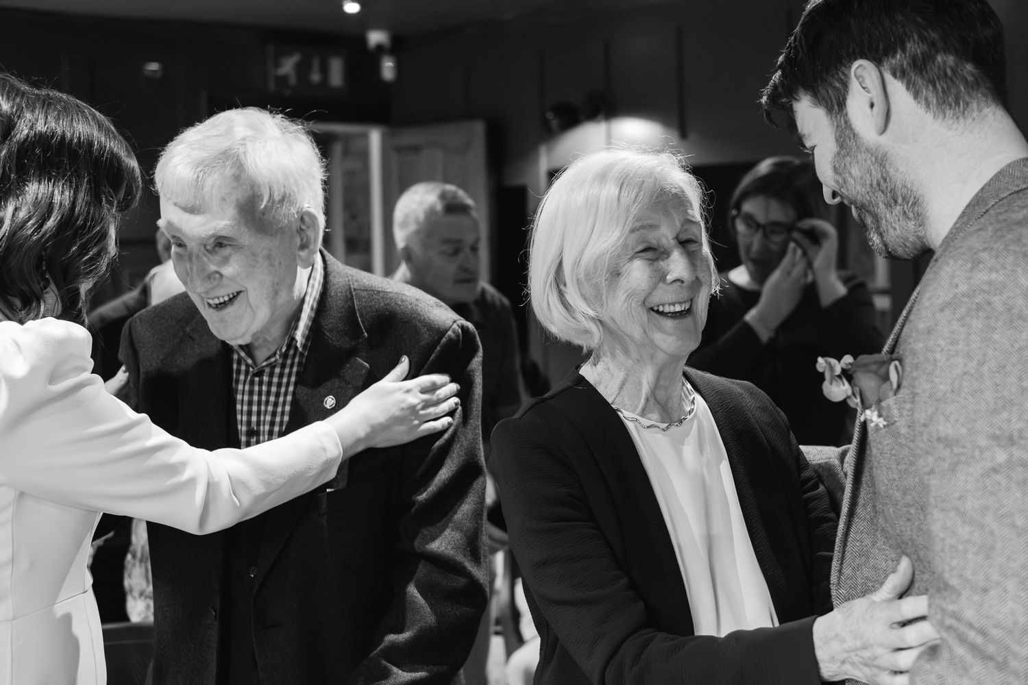 Black and white photo of elderly couple smiling and greeting guests at a social gathering event.