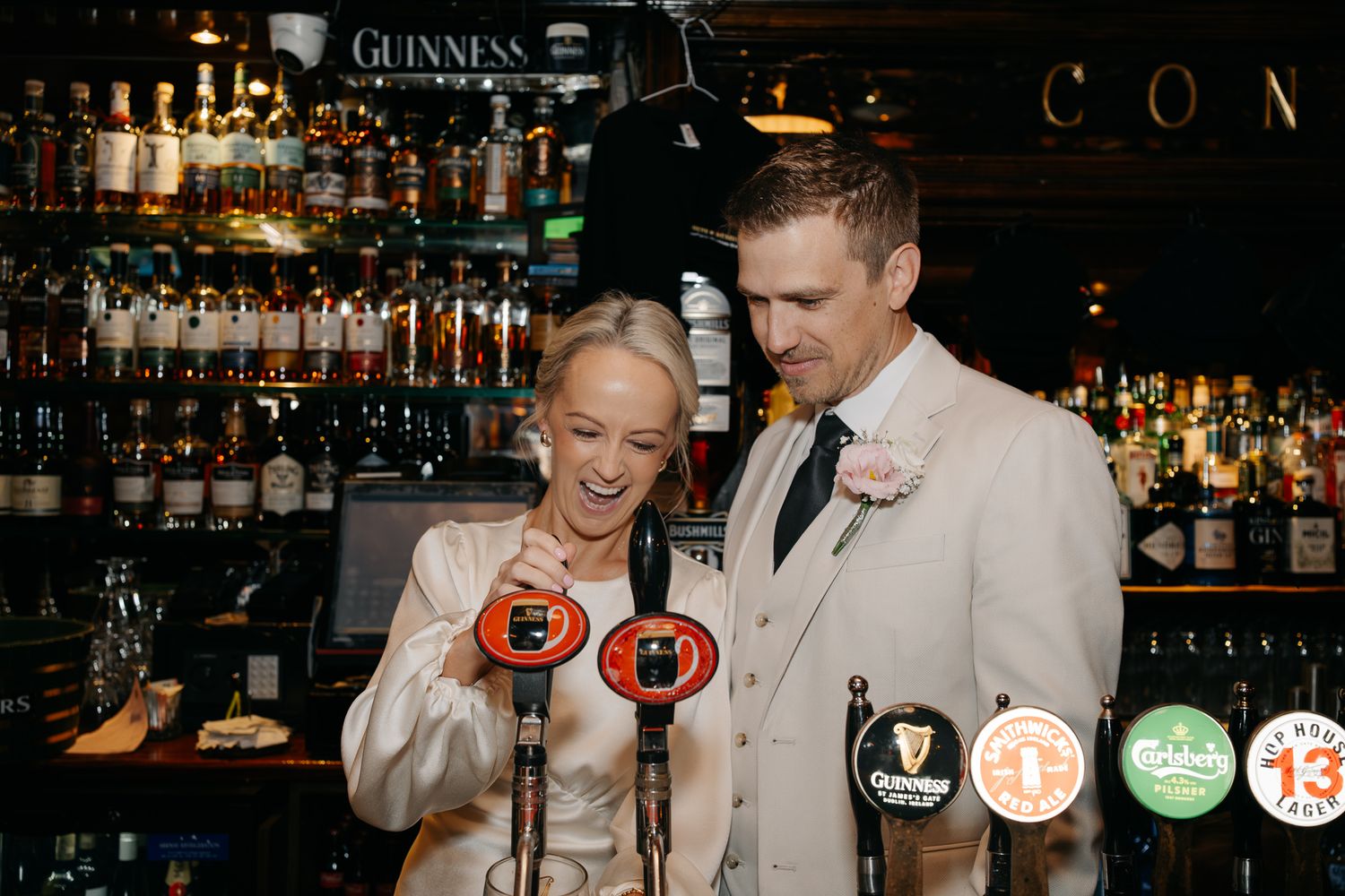 Bride and groom laughing while pulling Guinness taps at an Irish pub bar on their wedding day.