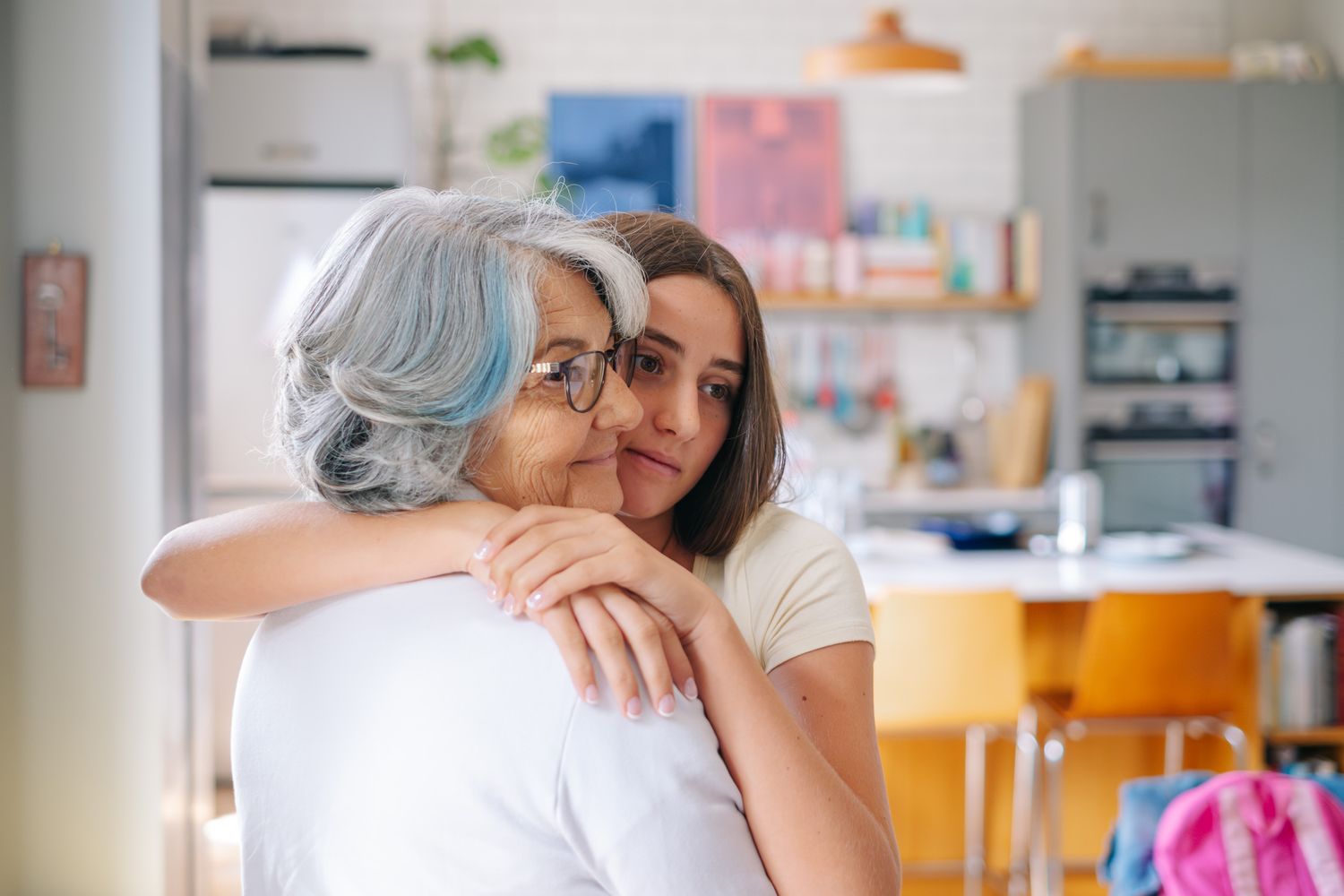 Elderly woman with gray hair hugging a young brunette woman warmly in a cozy home kitchen setting.