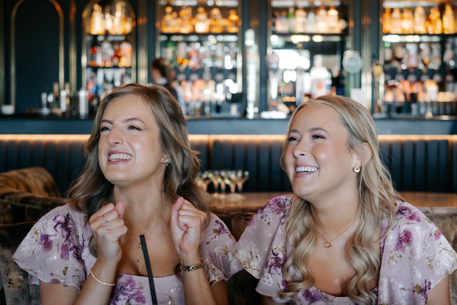 Two smiling women in floral dresses laughing together at a stylish bar with bottle-lined shelves in the background.