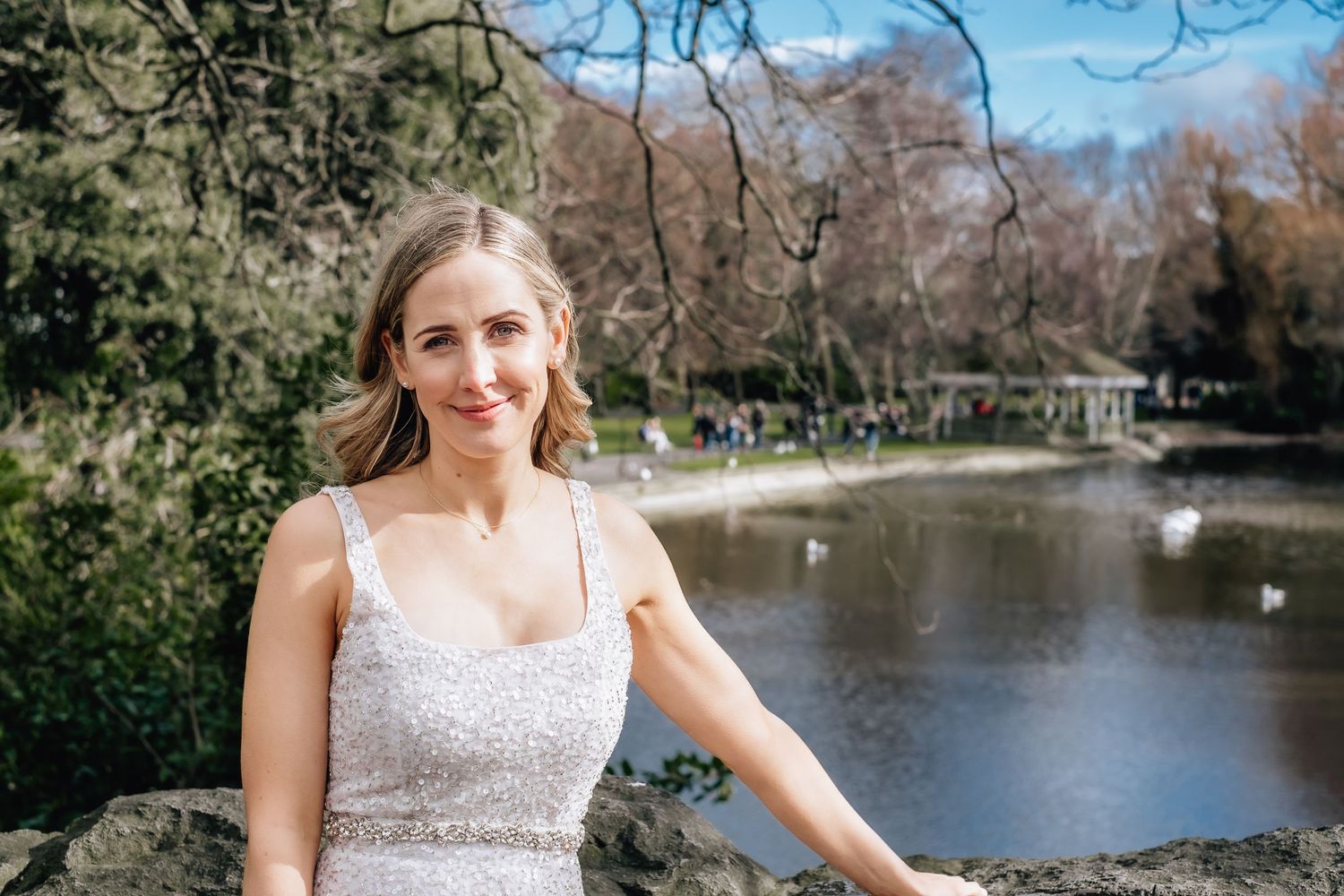 Bride in white dress poses by a scenic pond in a park setting.