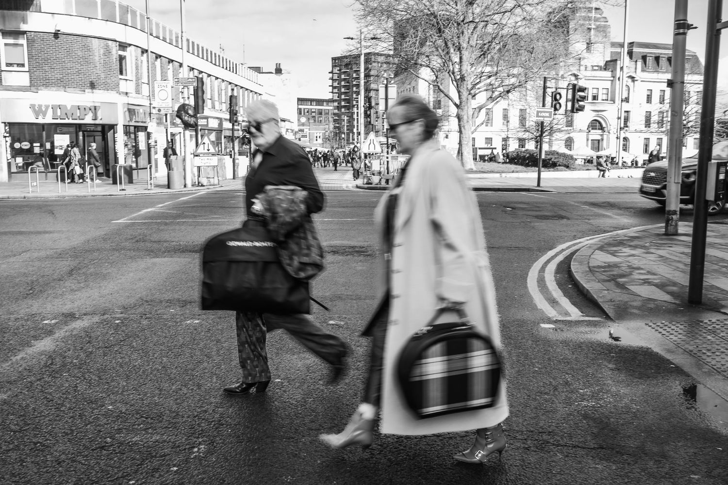 Black and white street scene with people crossing road carrying shopping bags.
