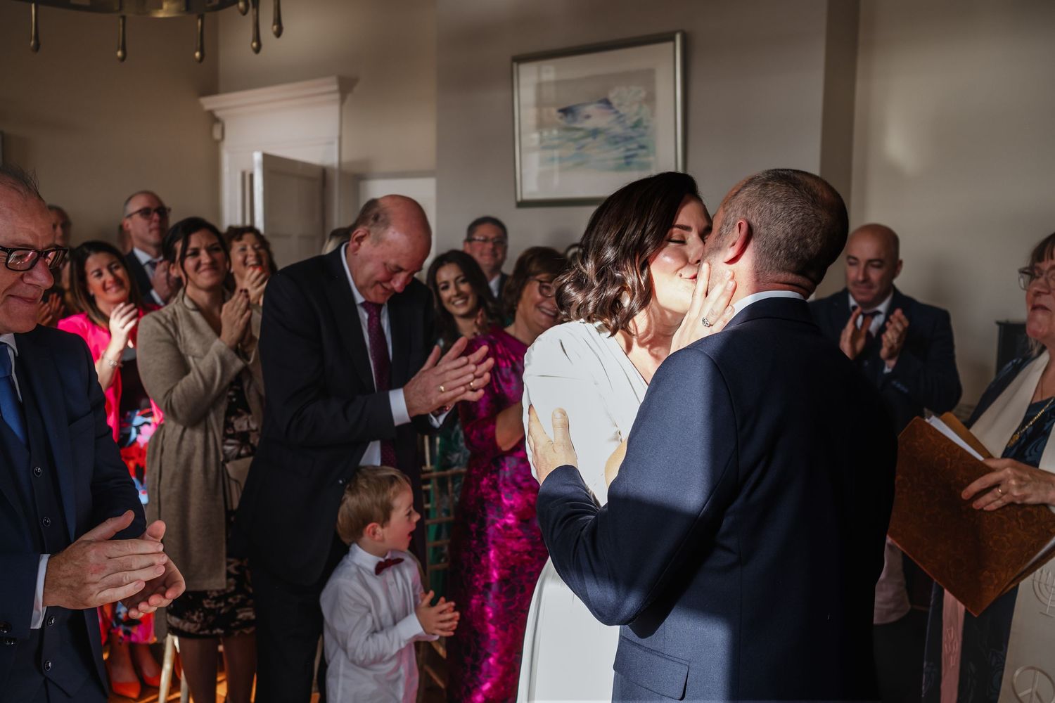Wedding guests applaud as couple shares kiss during intimate indoor ceremony celebration.