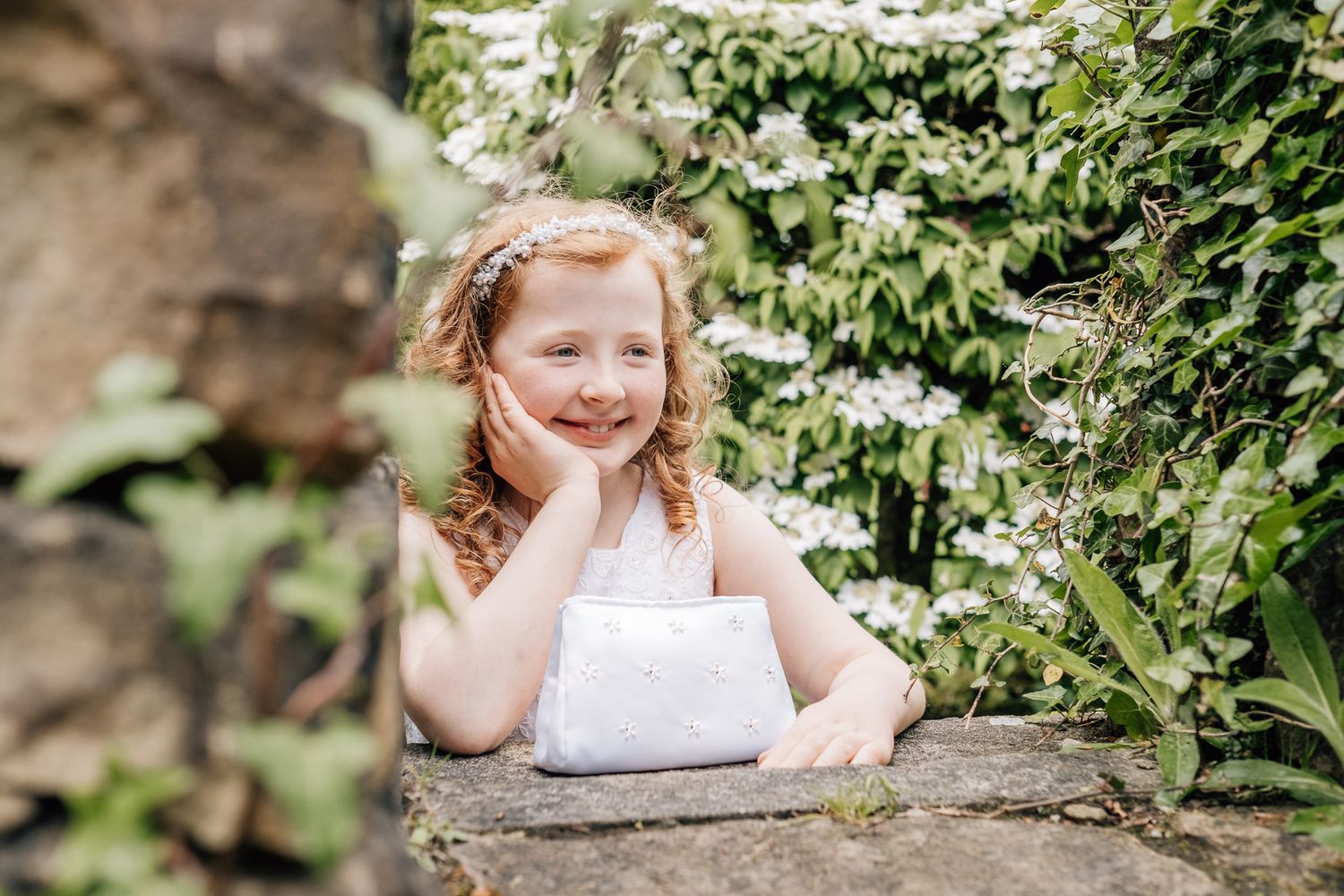 A smiling red-haired girl in a white dress poses among lush green foliage and white flowers in a garden.