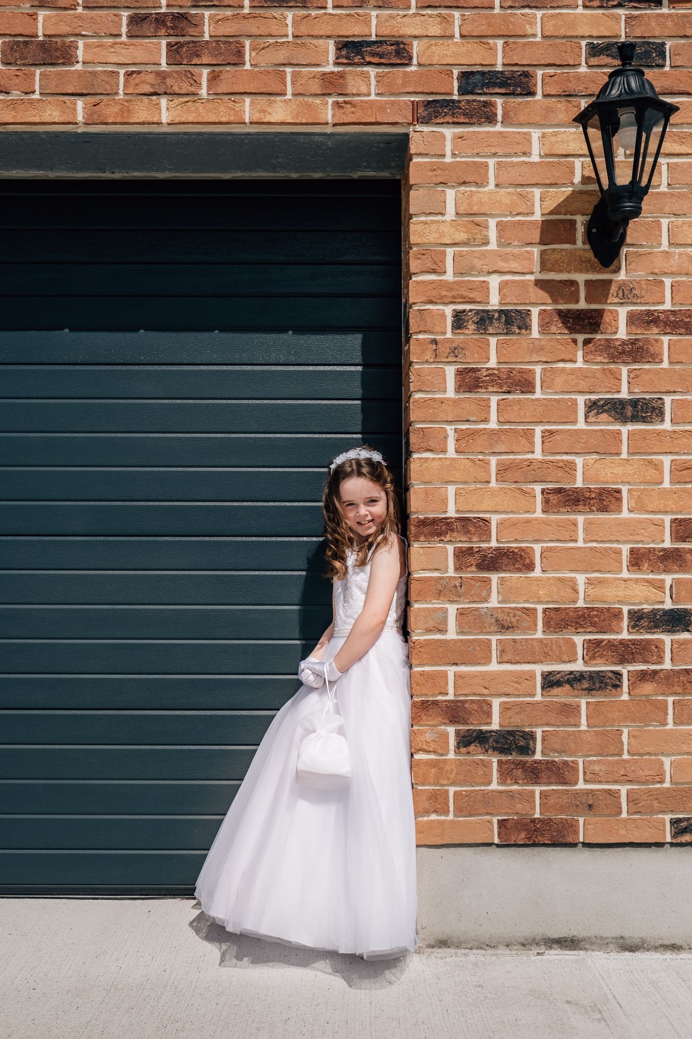 Young girl in white First Communion dress and gloves poses smiling beside brick wall and garage door.