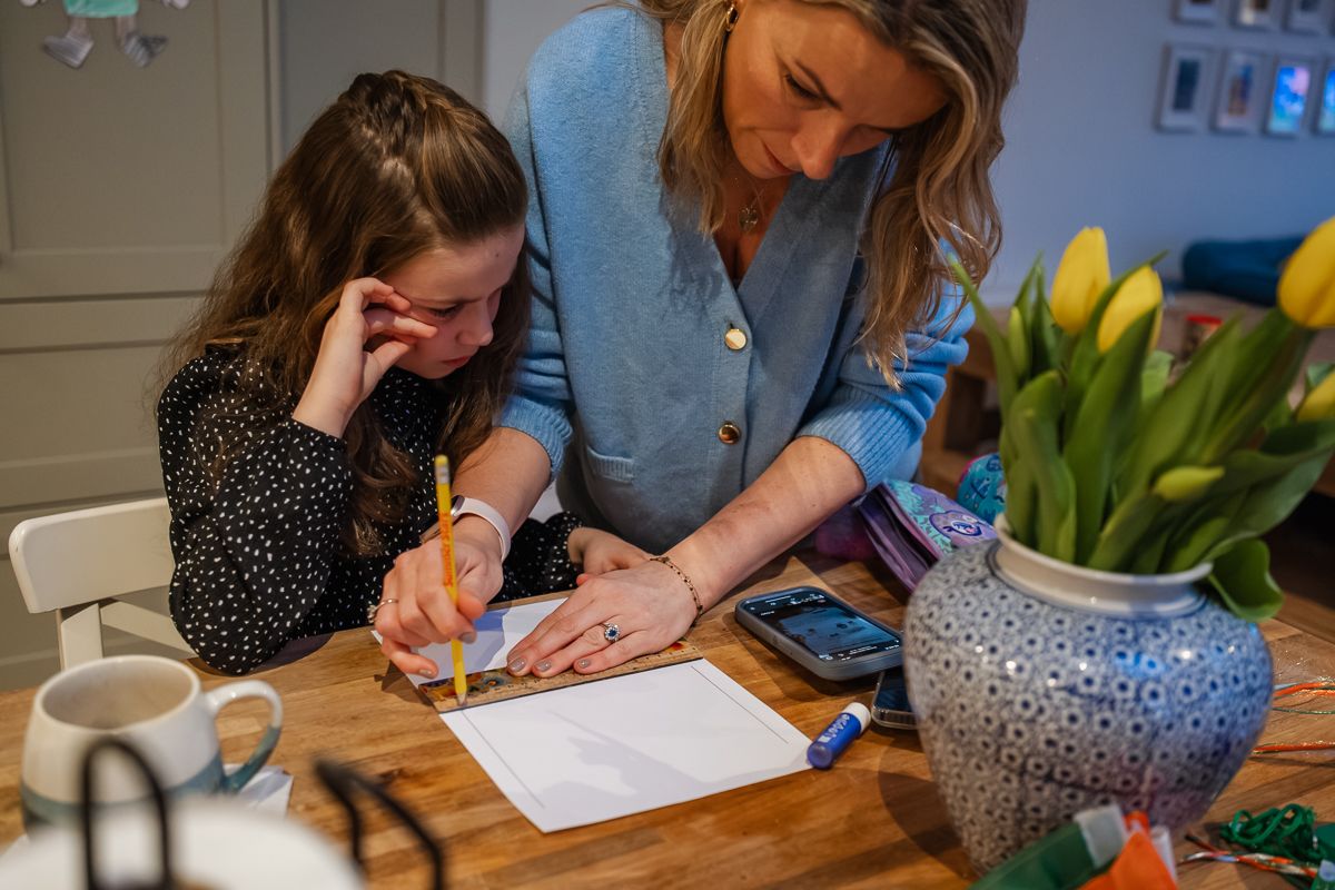 Mother helping young daughter with homework at wooden table with yellow tulips in a blue vase nearby.