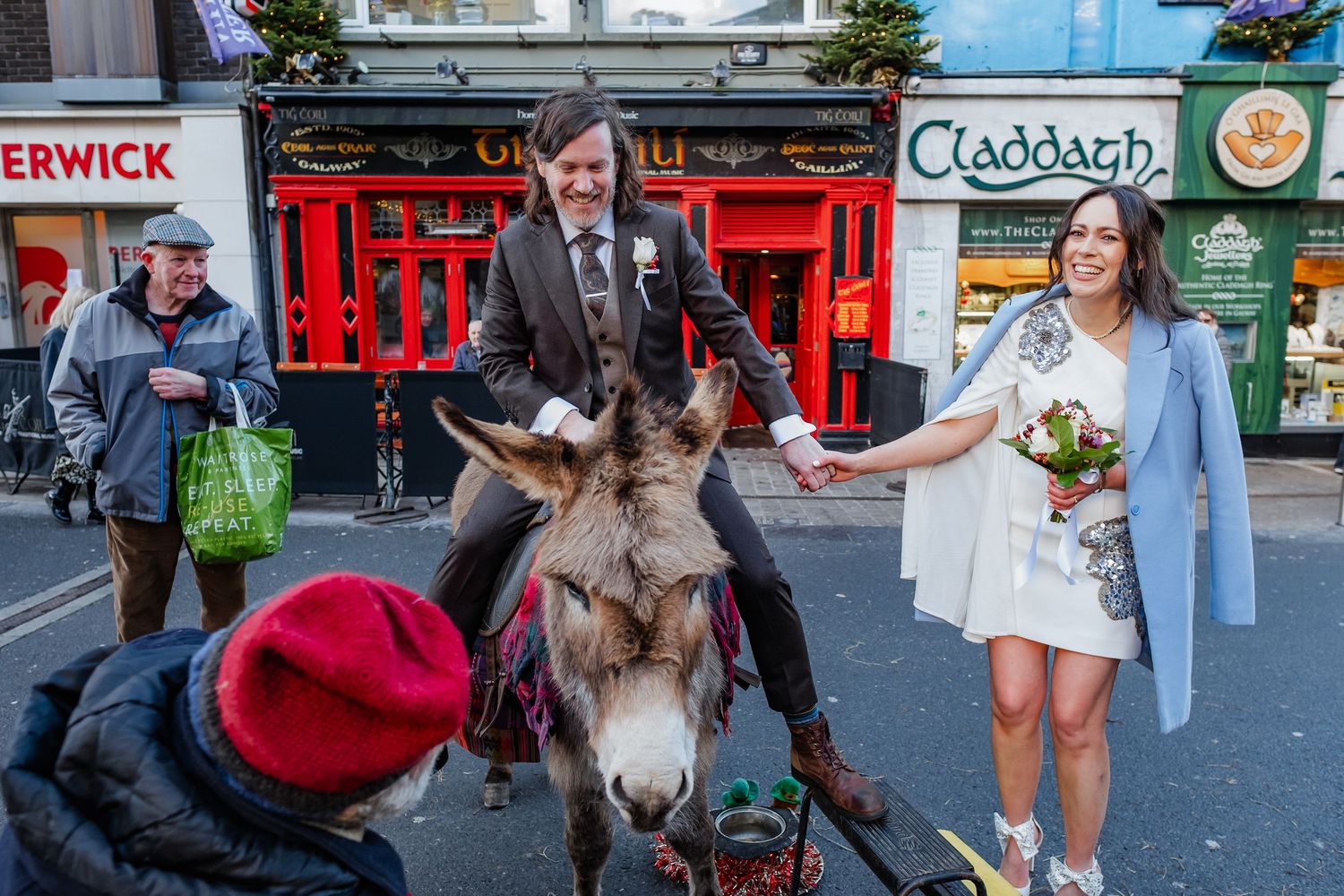 Groom rides donkey while bride holds bouquet on colorful Irish street near Claddagh pub during wedding celebration.