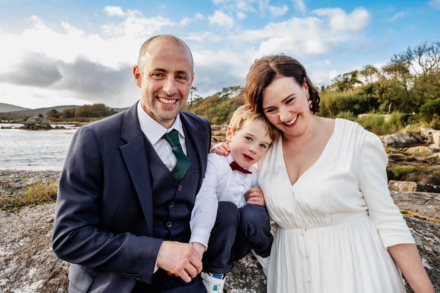 Happy family in wedding attire smiling outdoors by a scenic loch, with groom in navy suit and bride in white dress.