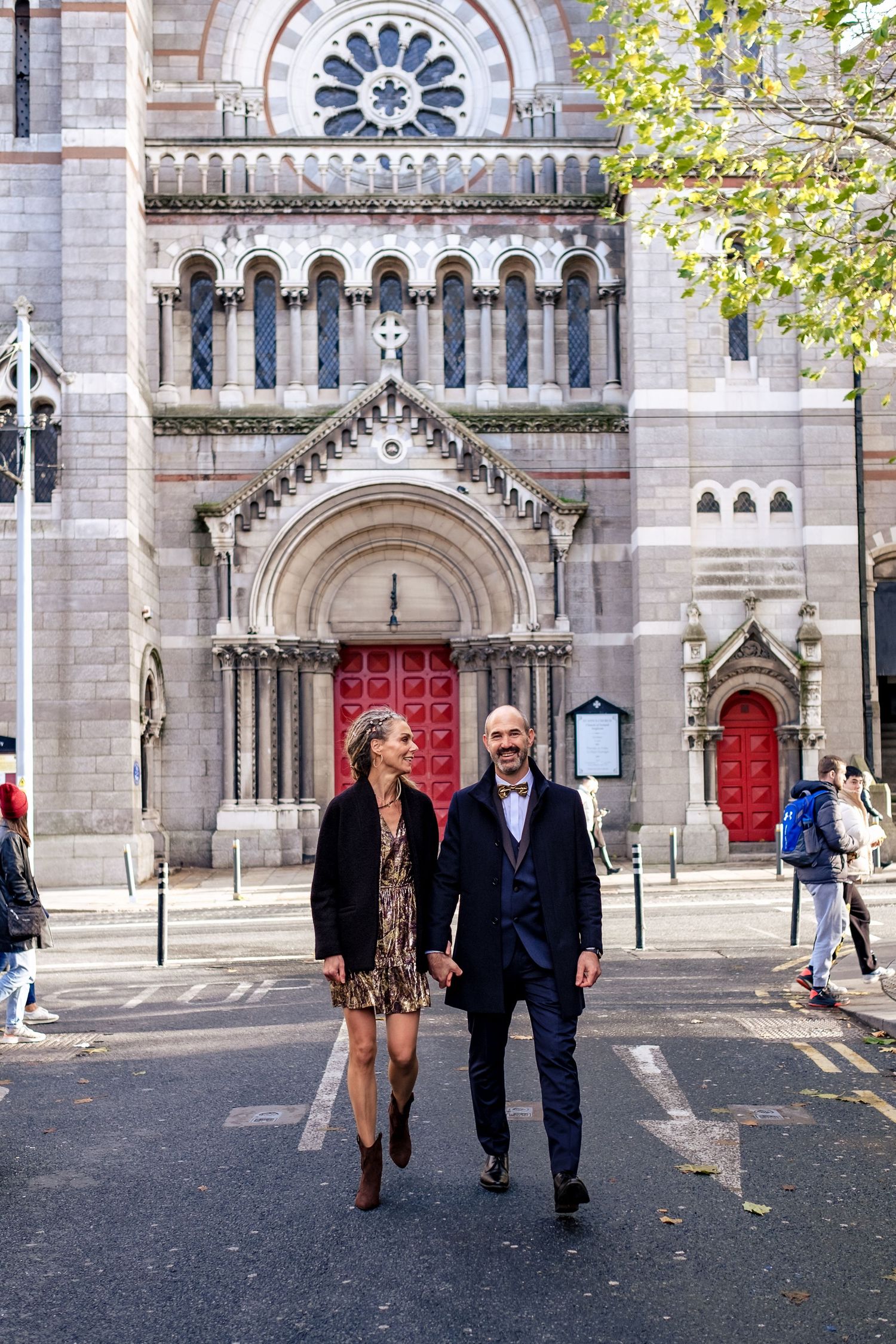 Couple holding hands walking in front of a grand stone church with red doors in a city street.