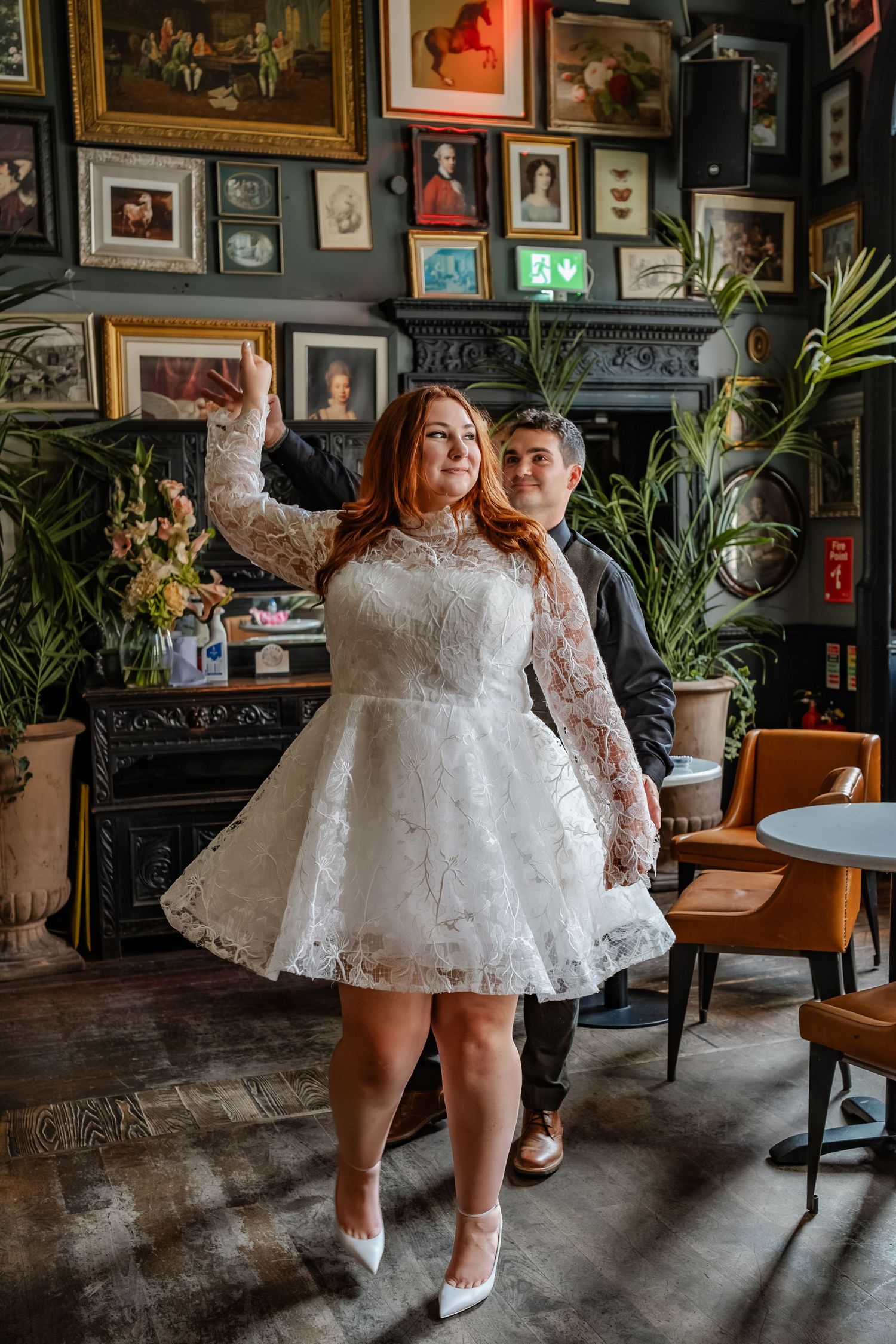 Person in white lace dress posing against wall of framed artwork in vintage pub setting.