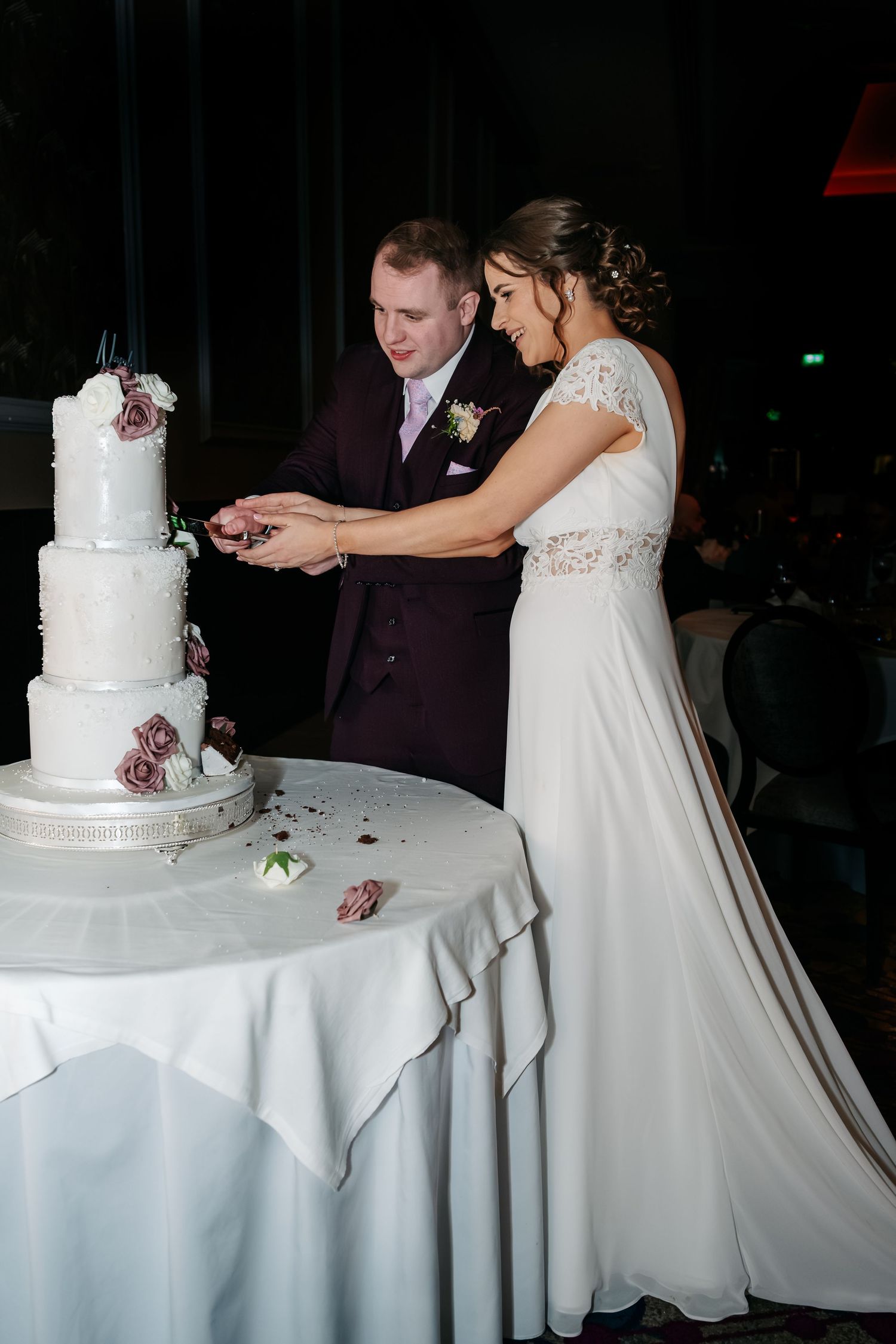 A couple in wedding attire cuts into their elegant white tiered wedding cake decorated with burgundy flowers.