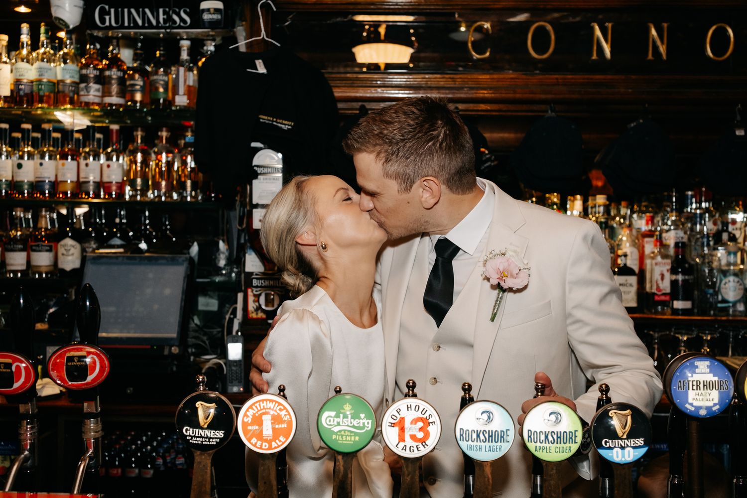 Newlywed couple sharing a romantic kiss at a pub bar with Guinness taps and bottles in the background.