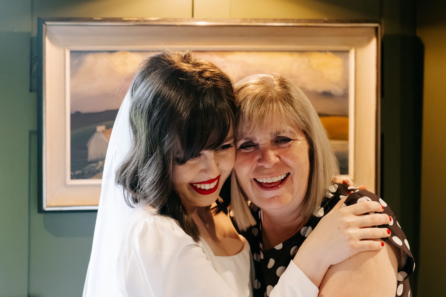 Two women sharing a warm hug and smiling, one wearing a white dress and veil, in front of a framed painting.