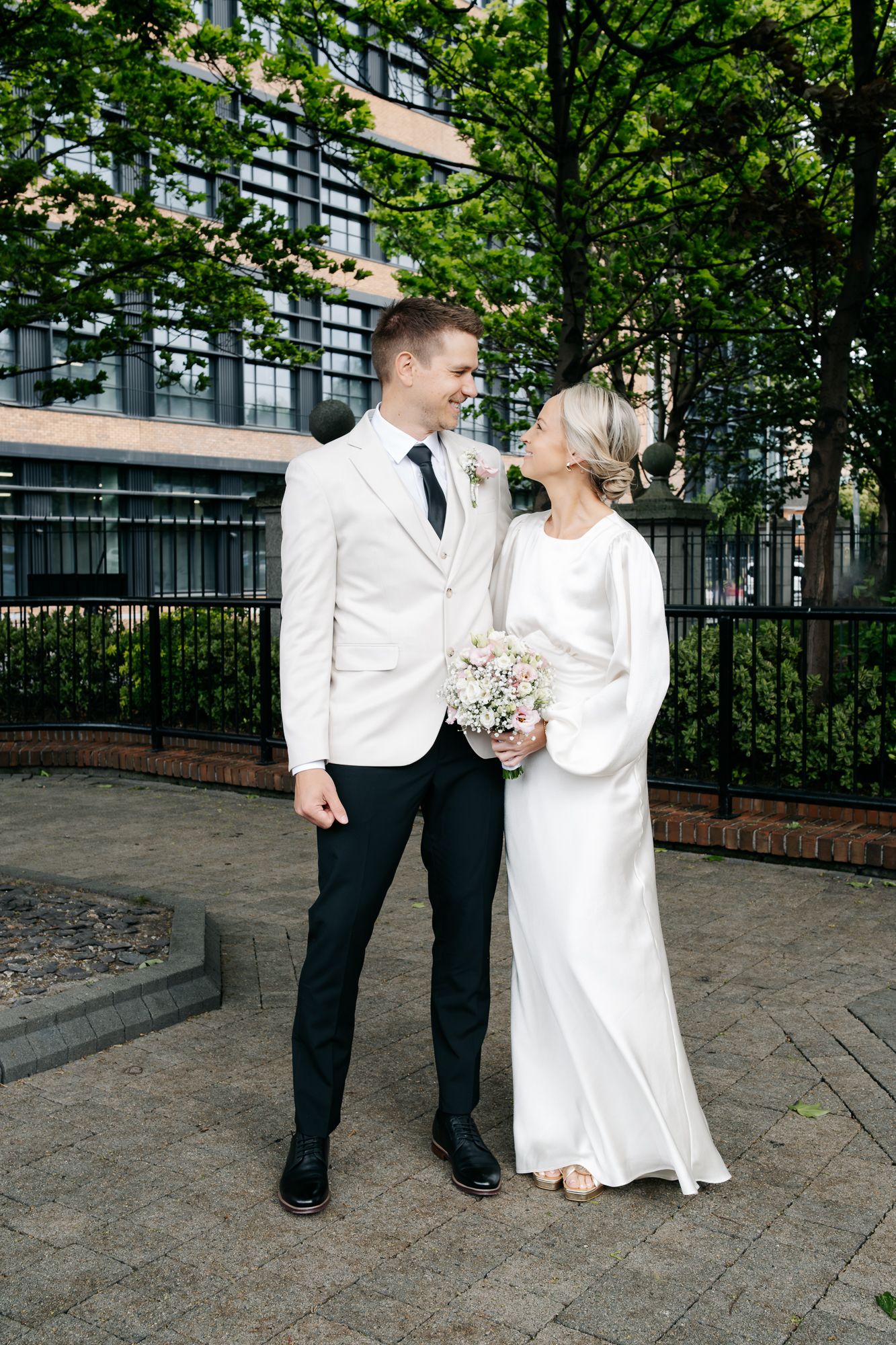 Bride and groom in white wedding attire share a loving glance outdoors, holding a pastel bouquet near a modern building.