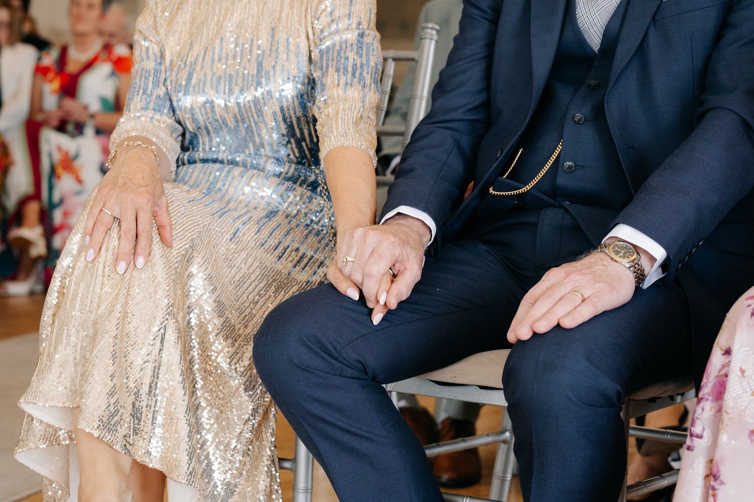 Couple holding hands at wedding ceremony, woman in sequin dress, man in navy suit with gold watch.