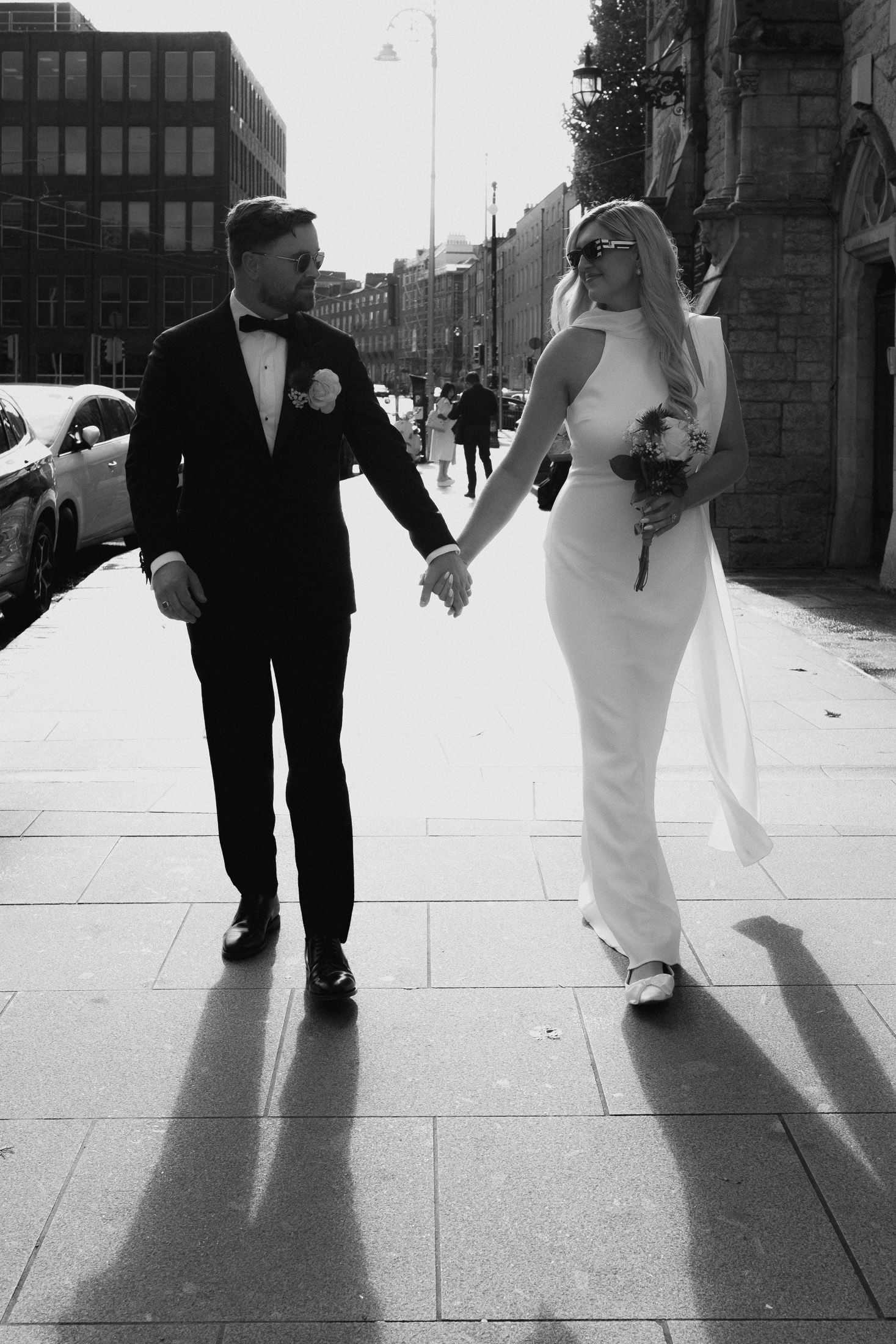 Black and white photo of a bride and groom holding hands walking on a city street, bride in white gown, groom in tuxedo.