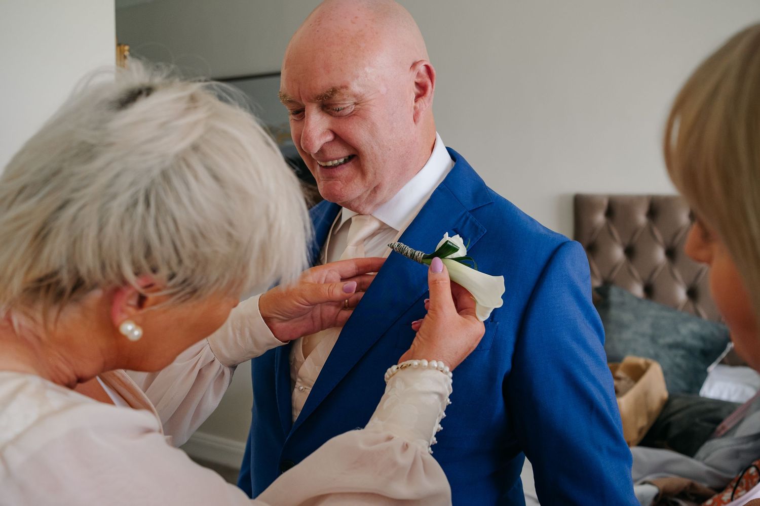 Women helping bald groom pin boutonniere on blue suit jacket before wedding ceremony.