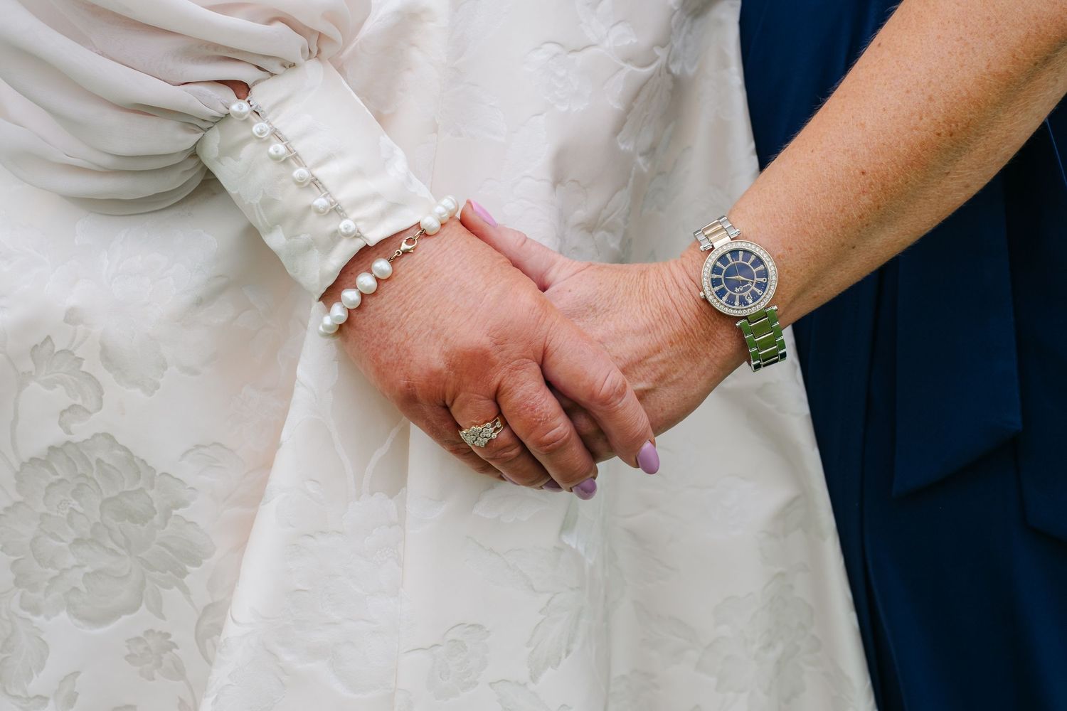 Close-up of bride and guest holding hands, showing pearl bracelet and blue-faced watch.