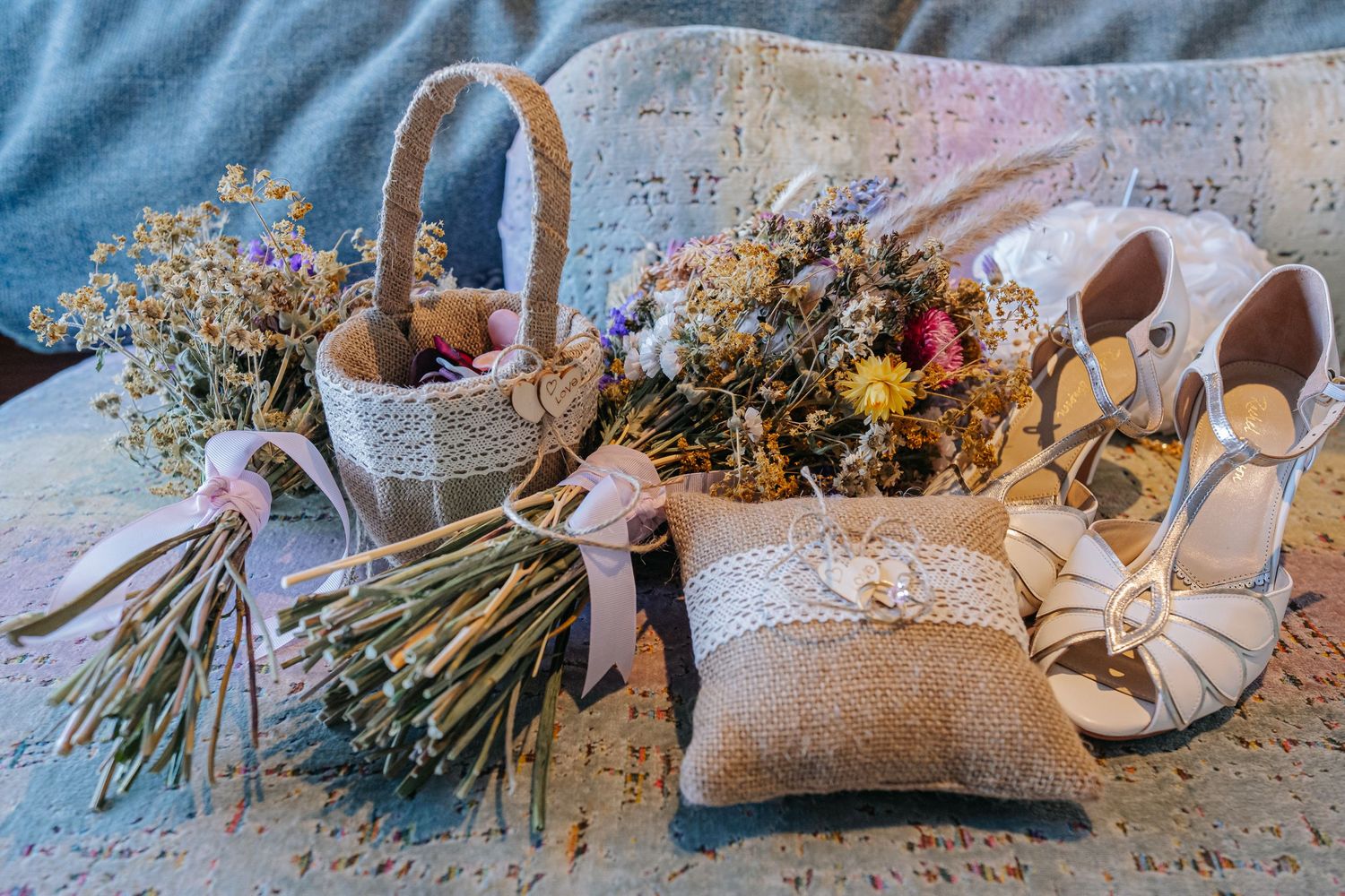 Rustic wedding flat lay with dried flower bouquets, burlap basket, ring pillow, and white bridal heels on lace.