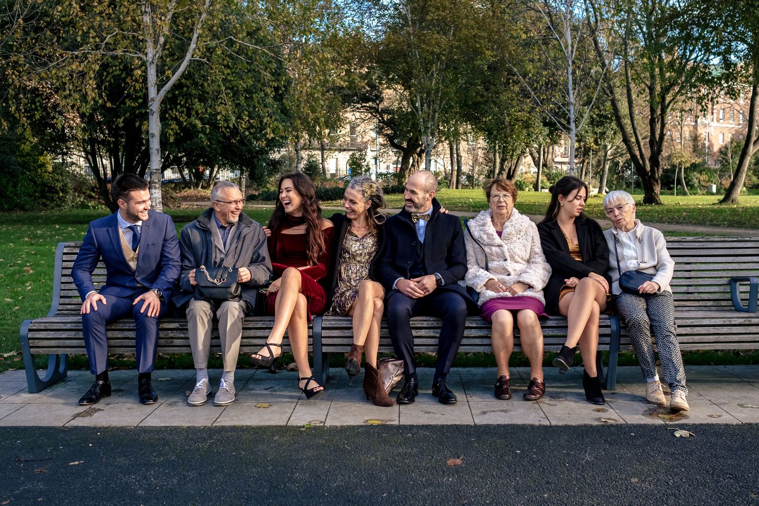 Eight formally dressed people sitting together on a park bench on a sunny day.