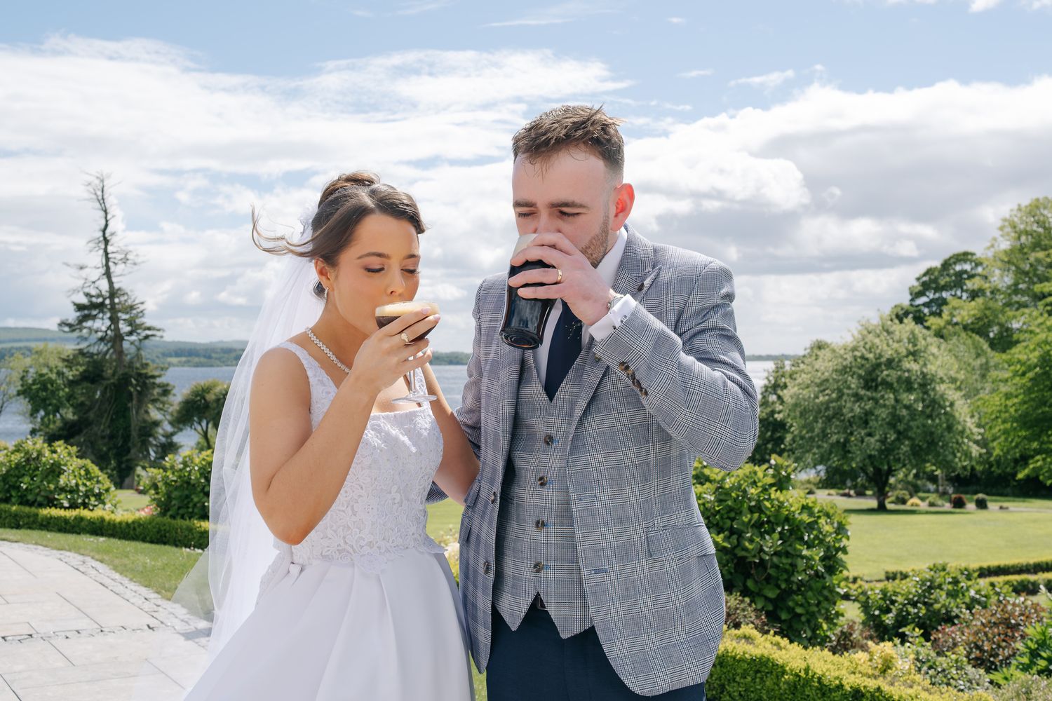 Bride and groom sipping drinks outdoors on their wedding day, with lush gardens and a scenic lake in the background.