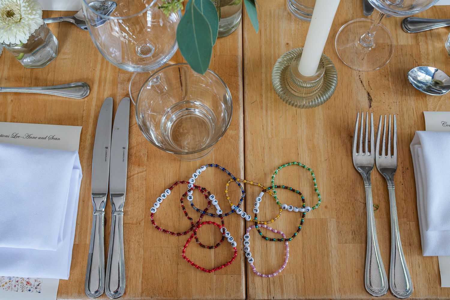 Elegant wedding table setting with wine glasses, silverware and colorful bracelets arranged on wooden surface.