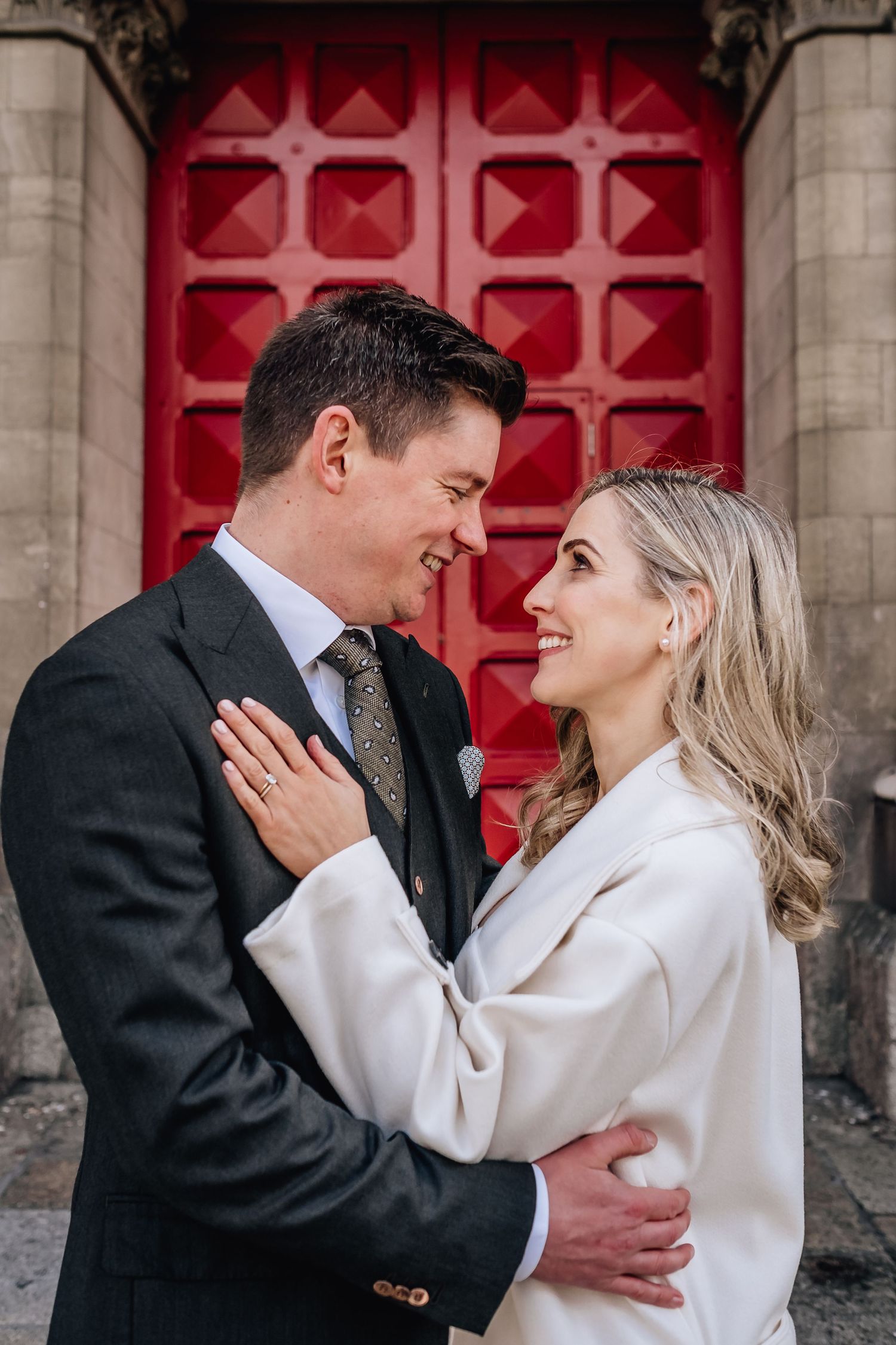 Couple shares intimate moment in front of ornate red church door during wedding photoshoot.