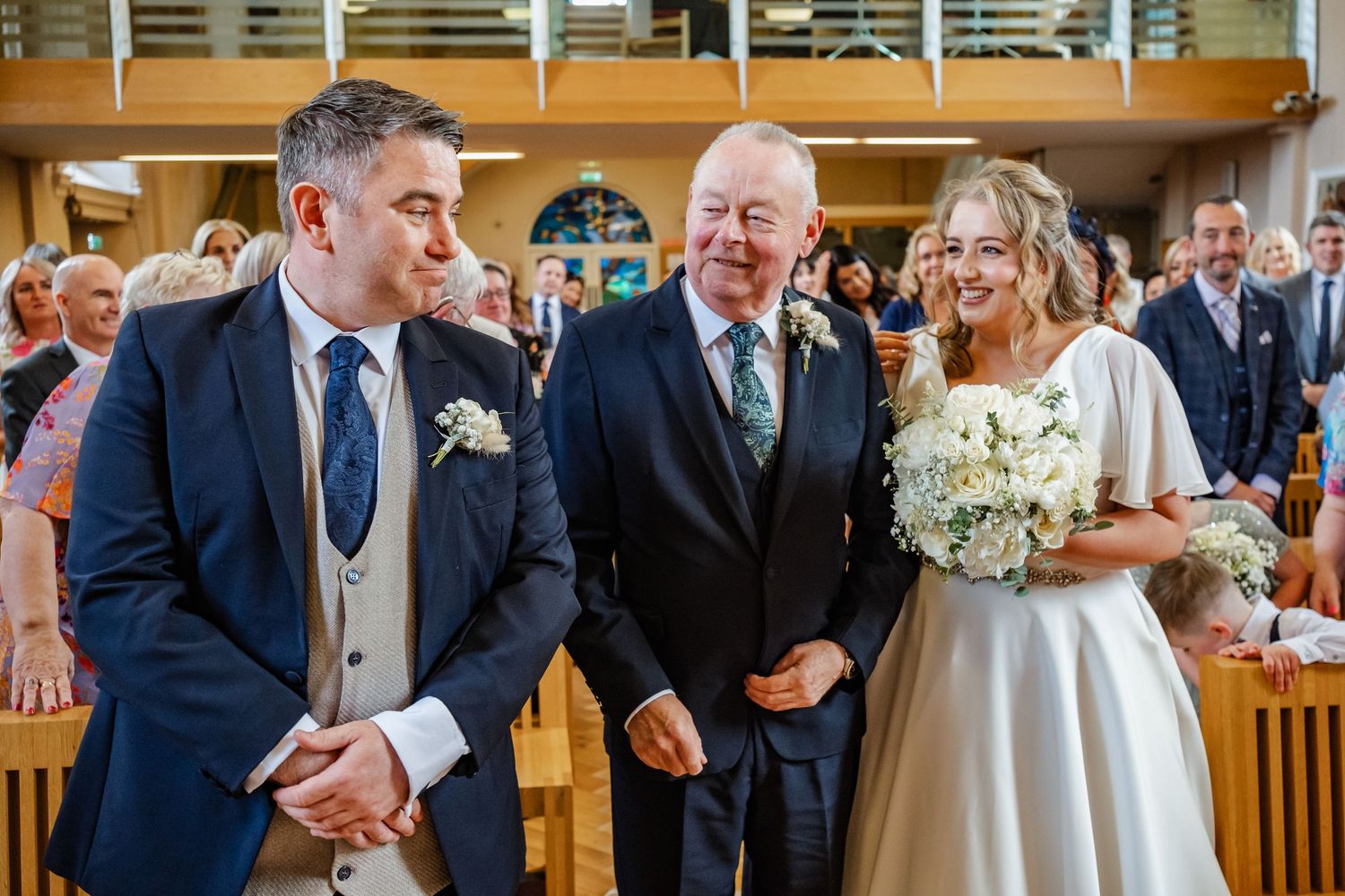 Wedding ceremony scene in bright church hall with wooden pews and balcony.