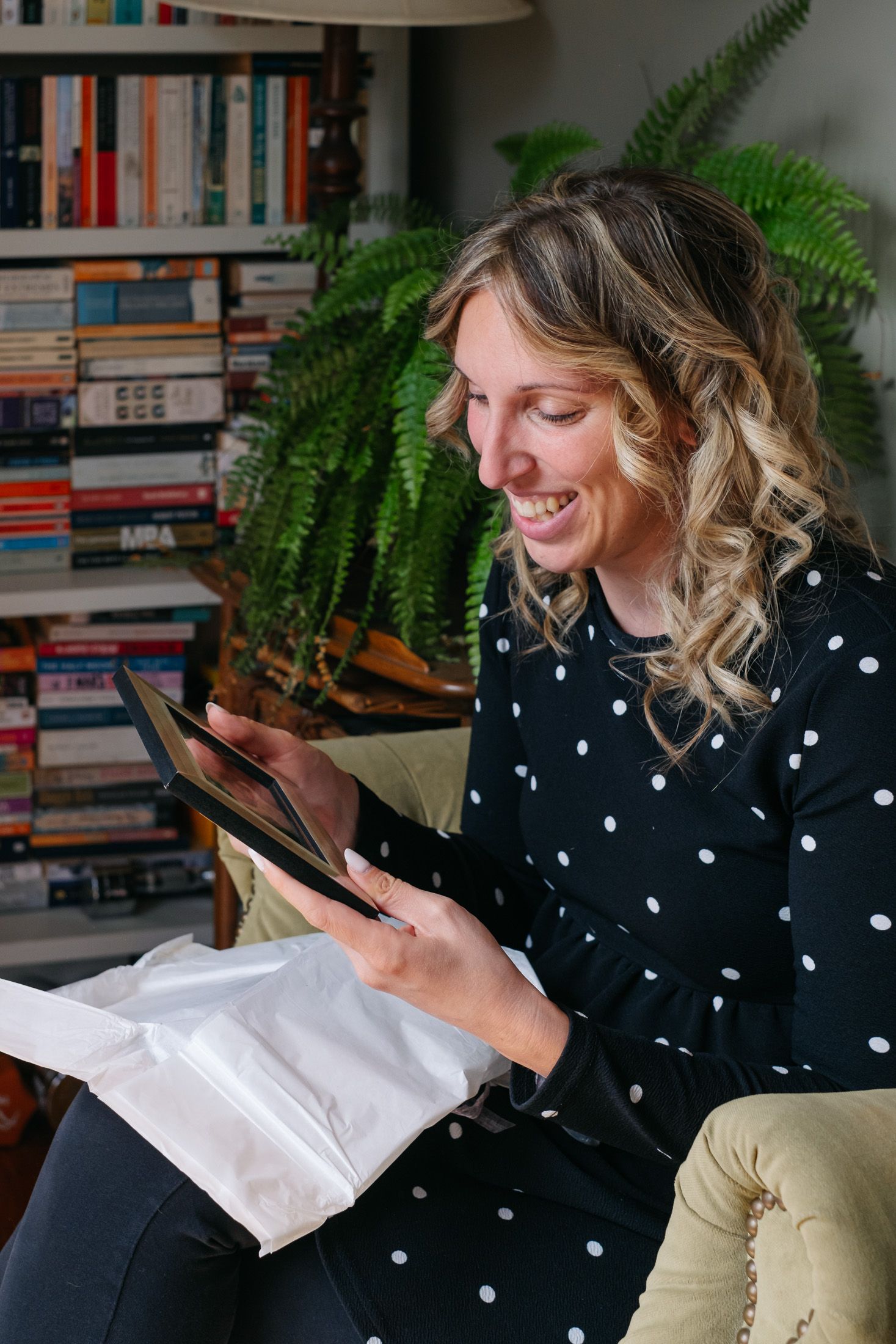 Smiling woman in polka dot blouse opening a package while seated in a cozy chair near a bookshelf.