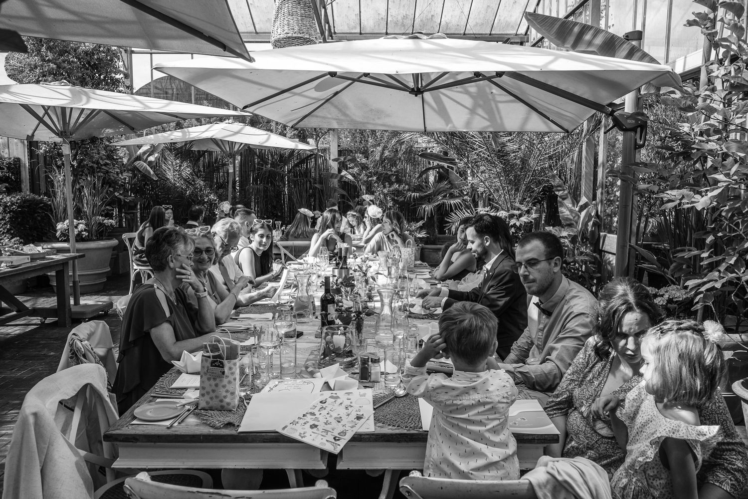 Black and white photo of a large group dining outdoors under white umbrellas at a lush garden restaurant.
