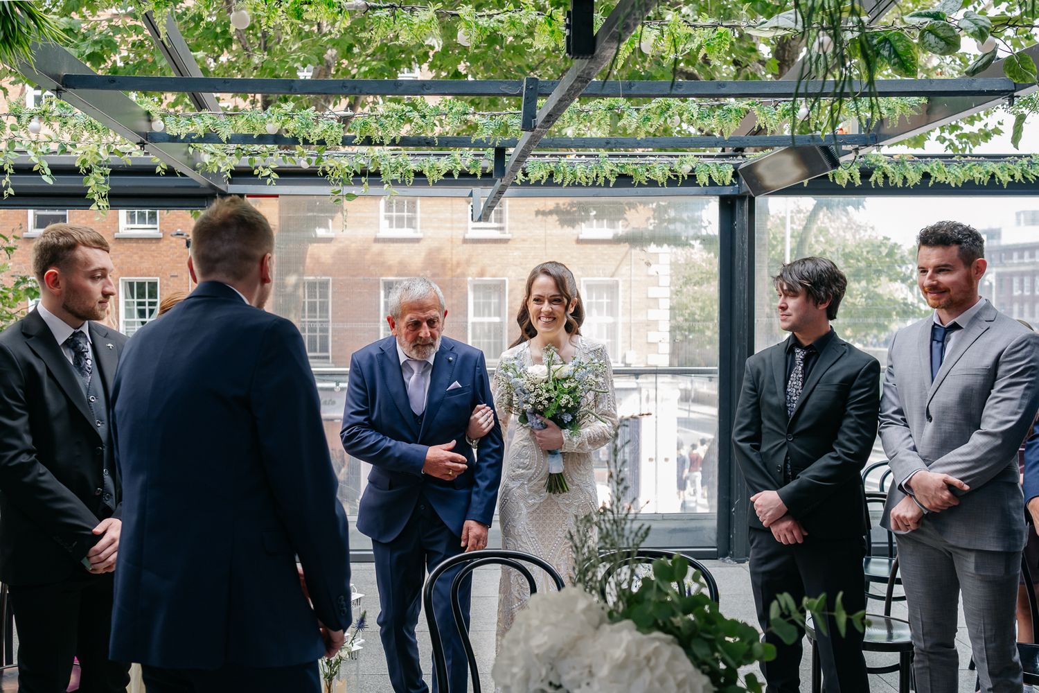 Wedding ceremony taking place under a greenery-covered pergola with guests in attendance.