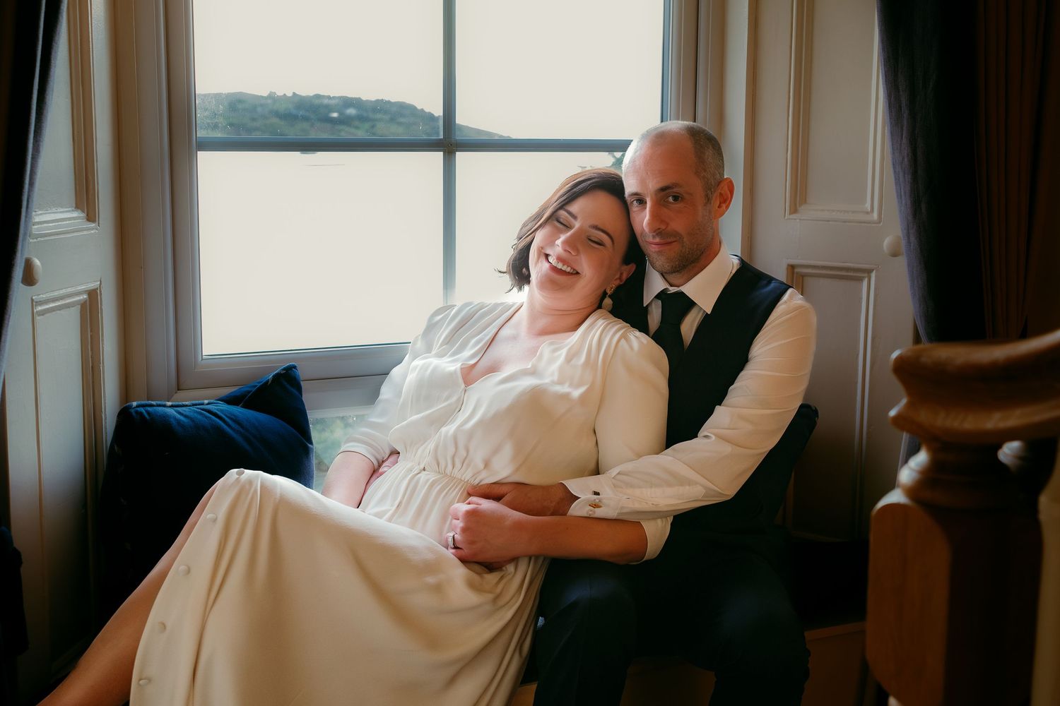 A couple shares a tender embrace by a window, the bride in a white gown laughing joyfully as the groom holds her close.