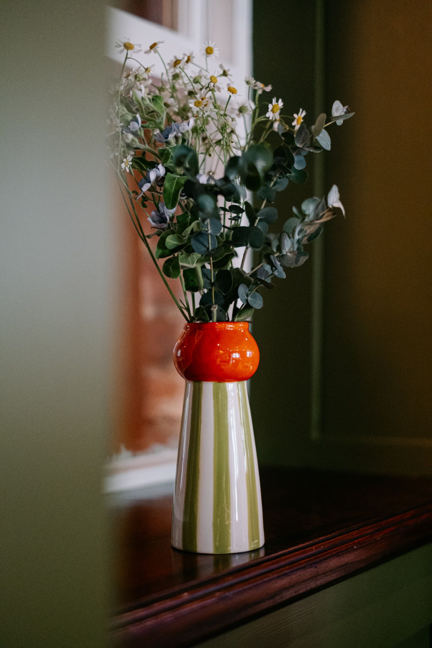 A striped green and orange ceramic vase holding eucalyptus and white wildflowers sits on a wooden shelf by a window.
