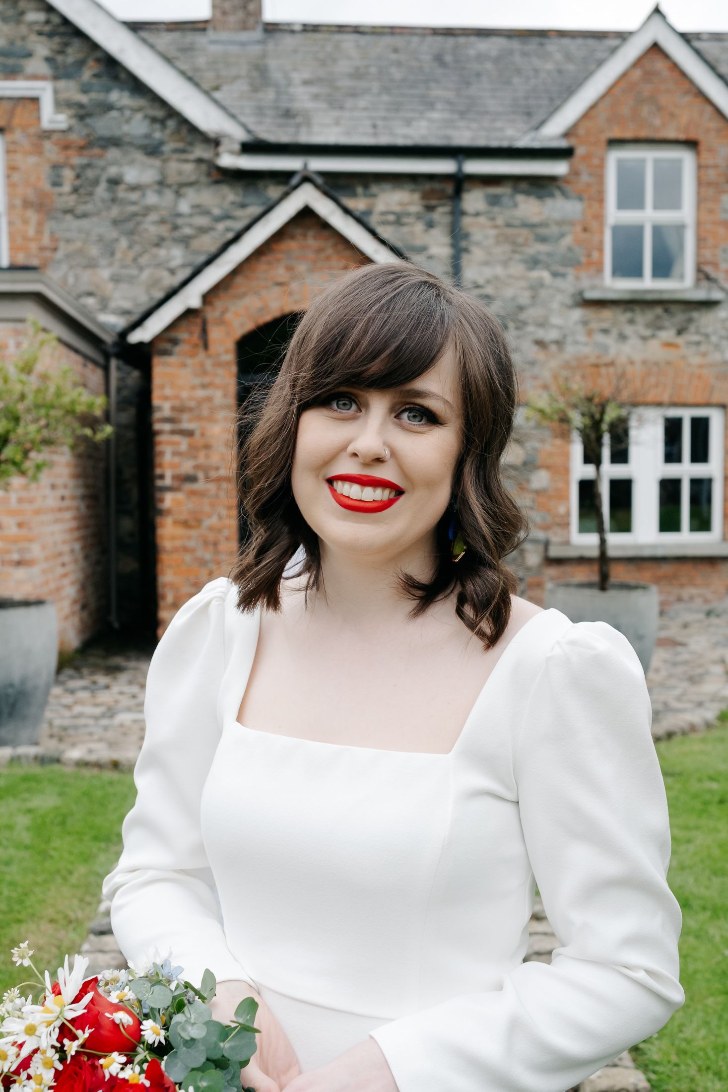 Smiling bride in white dress with red lips holds bouquet outside rustic brick venue.