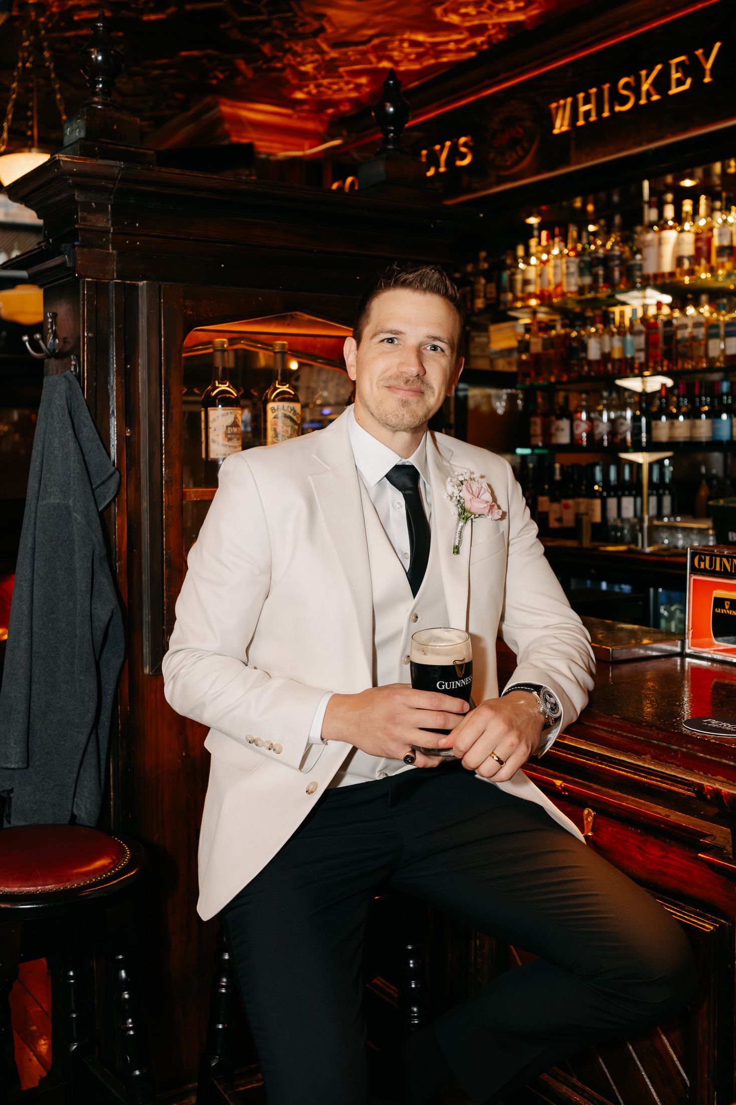 Groom in white blazer holding Guinness pint at ornate whiskey bar, wearing boutonniere and black tie.