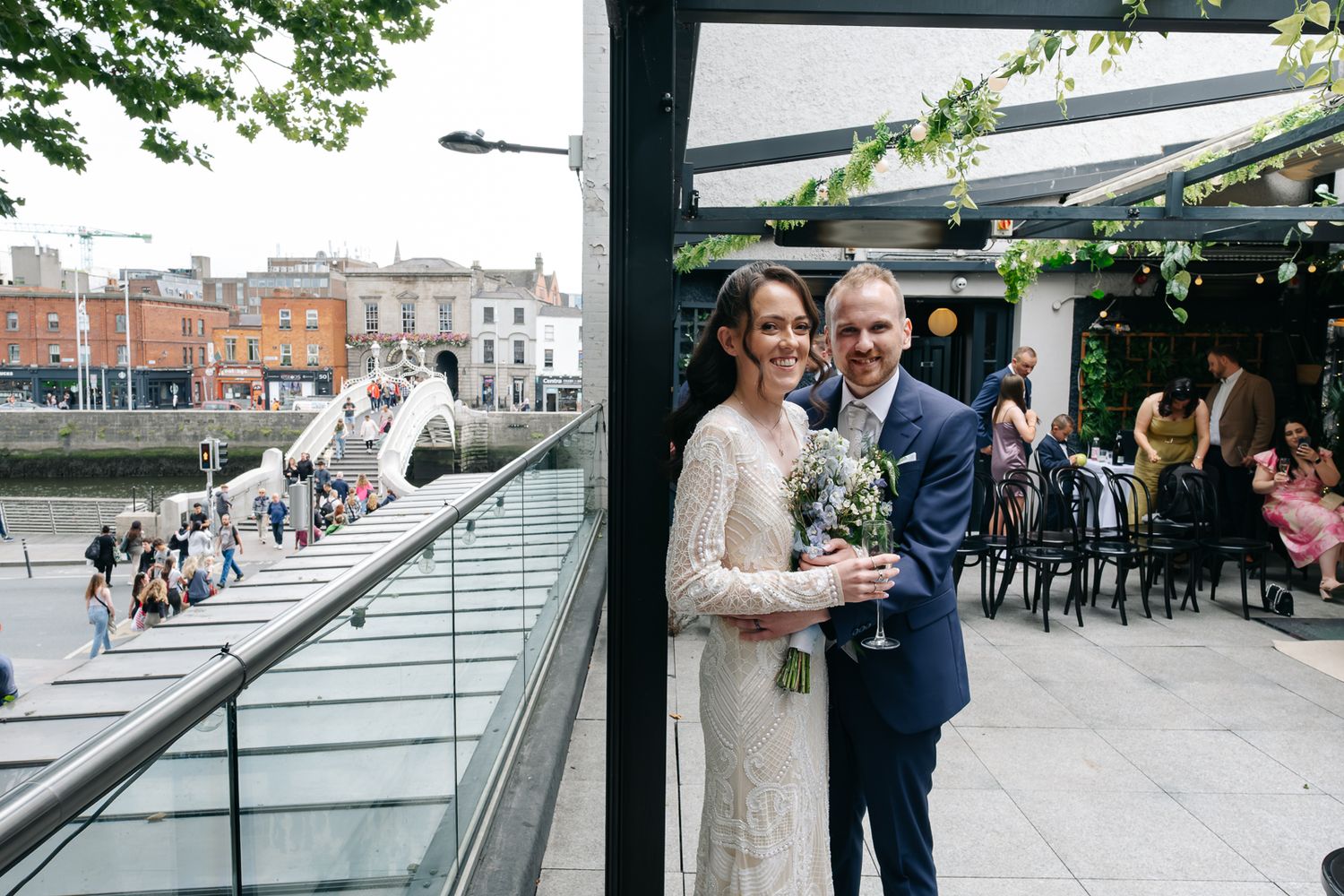 A couple poses together on a rooftop restaurant The Woollen Mills  Dublin with the Ha'penny Bridge and brick buildings in the background.