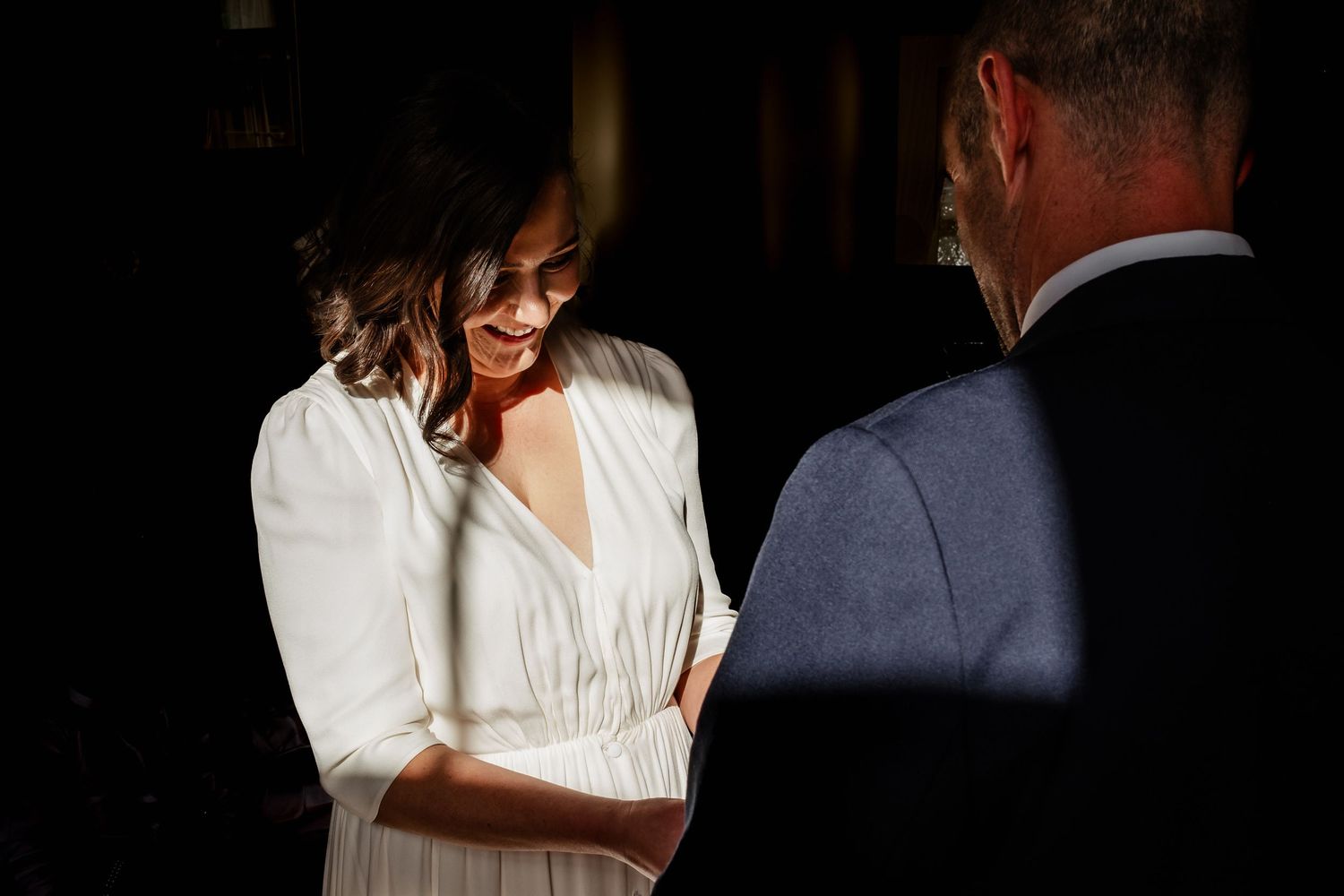A bride in a white dress smiles while holding hands with a groom in a blue suit during an intimate wedding ceremony.