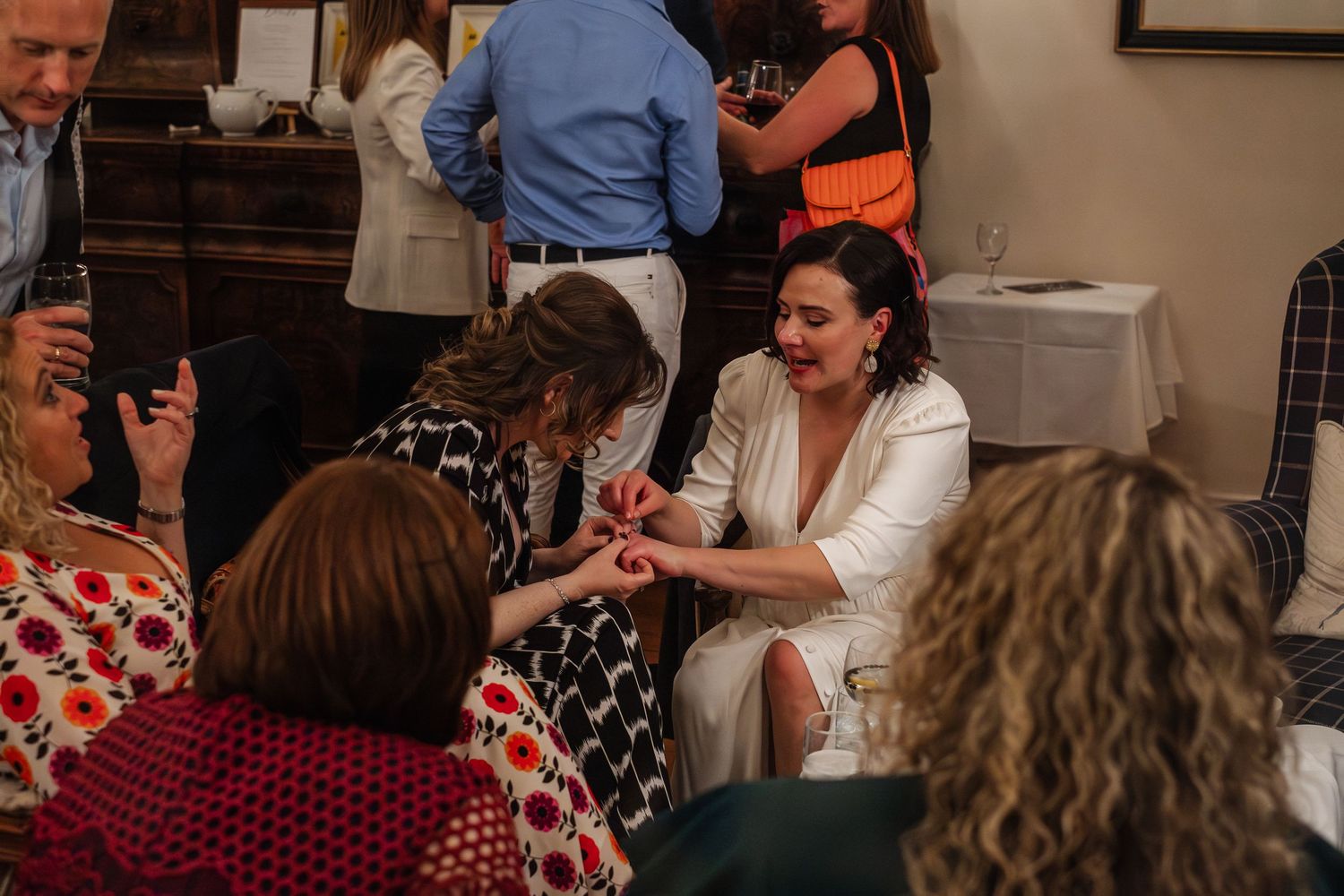 Bride in white dress showing her ring to seated wedding guests at indoor reception.