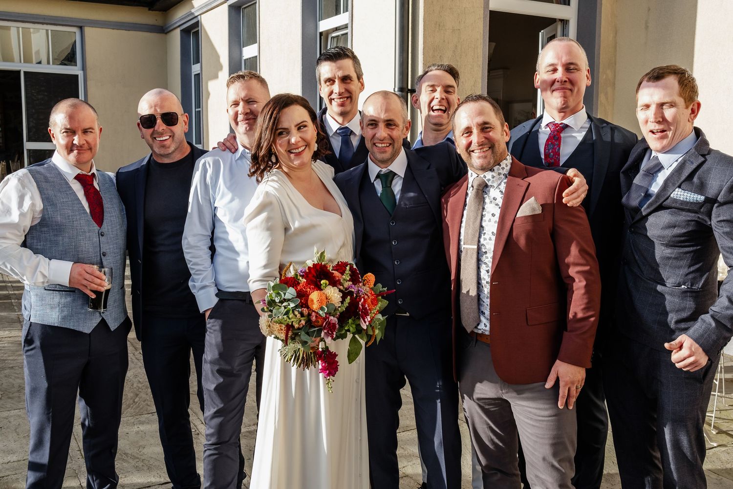 Bride holding colorful bouquet posing with group of groomsmen outside at wedding celebration.