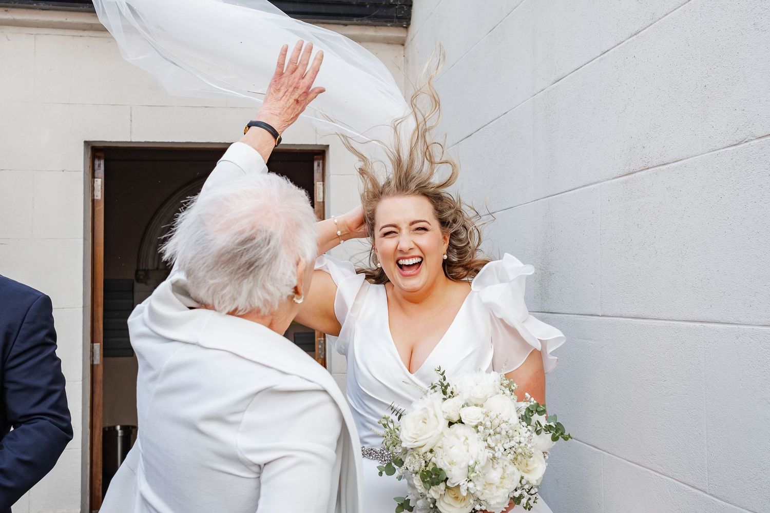 Laughing bride in white gown holds bouquet as elderly guest tosses veil outside church on wedding day.