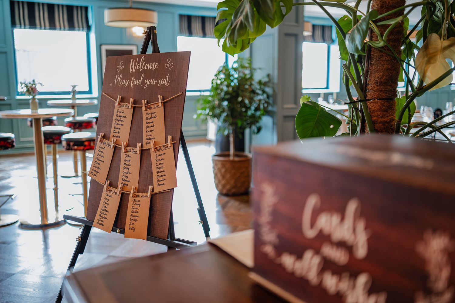 Wedding seating chart board displayed on an easel in a modern venue with teal walls and potted plants.