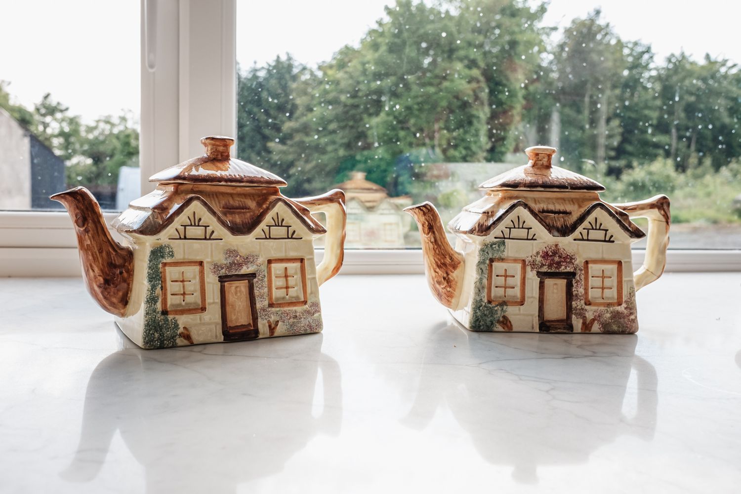 Two decorative ceramic teapots shaped like cottages sitting on a windowsill.