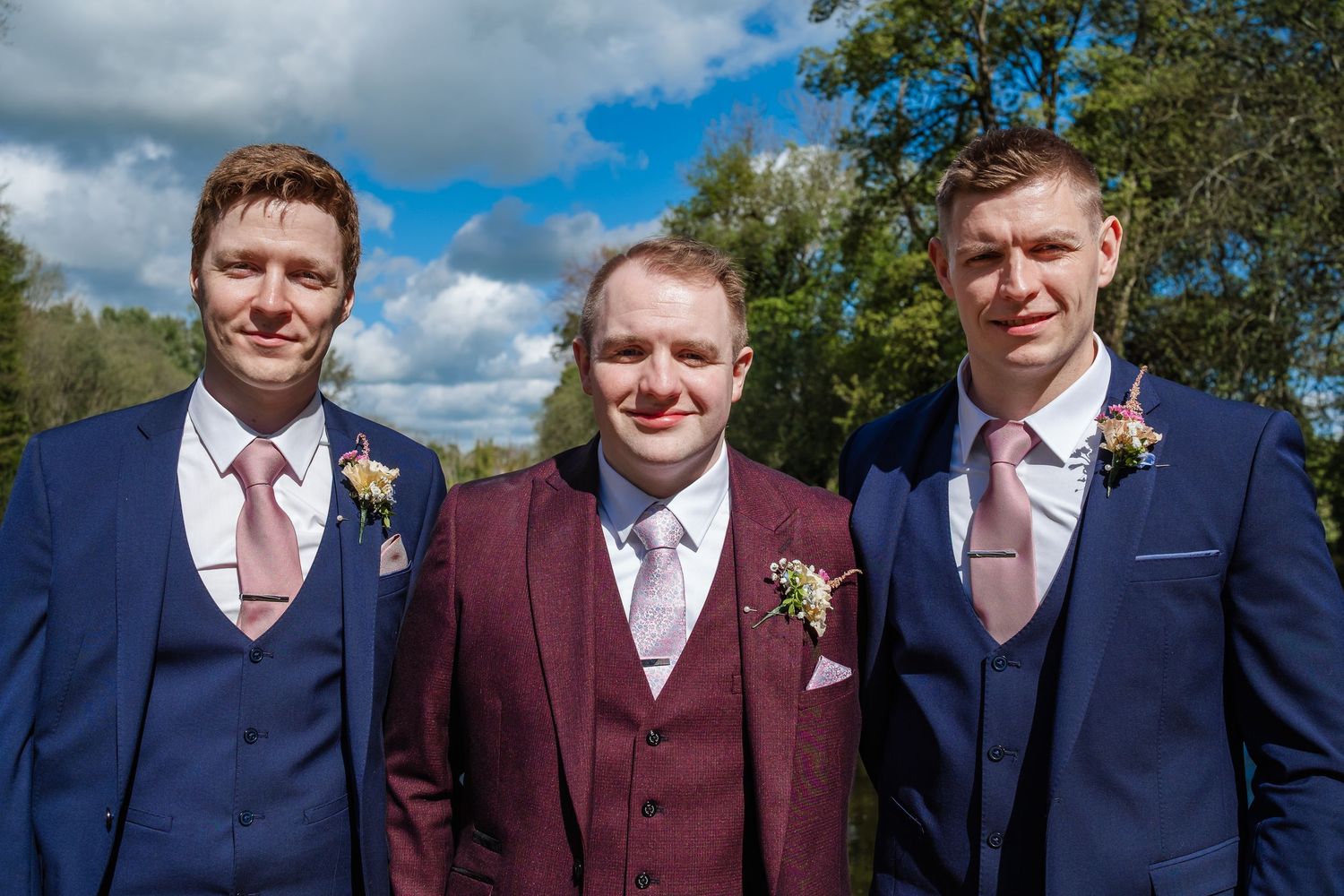 Groomsmen in blue suits and burgundy vests pose outdoors with fluffy white clouds in background.