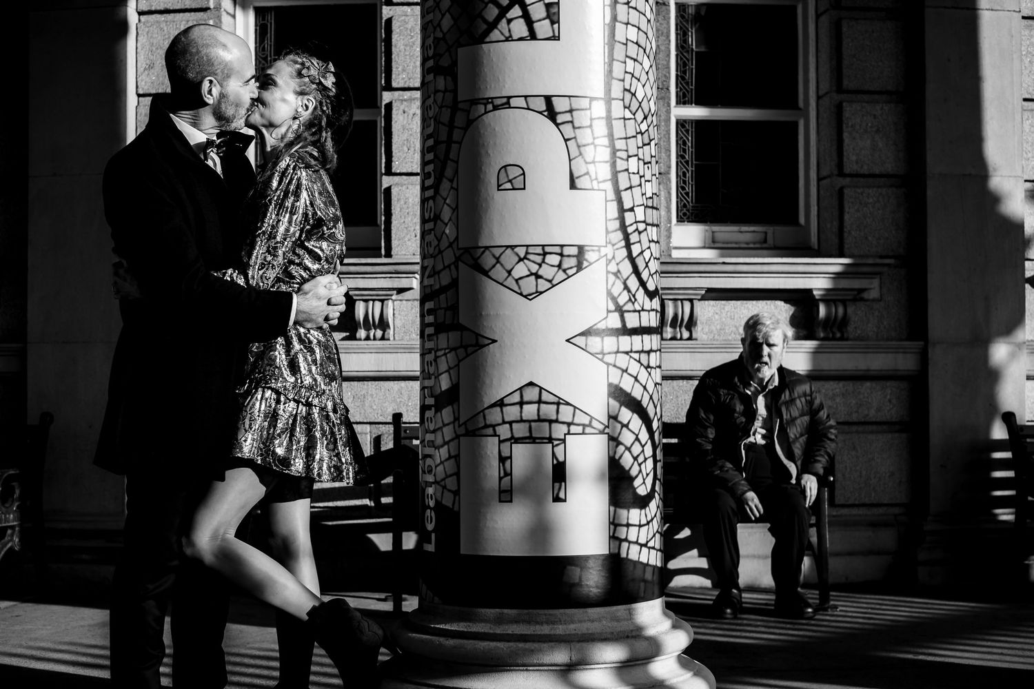 Black and white photo of a couple embracing near a decorative column while an elderly man sits watching in the background.