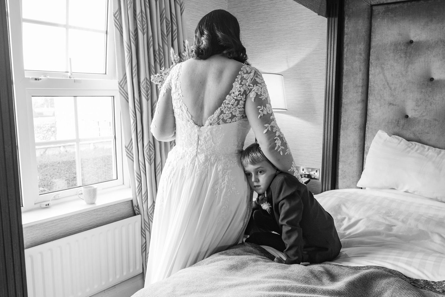 Black and white photo of a bride in lace gown standing by window with young boy in suit on bed beside her.