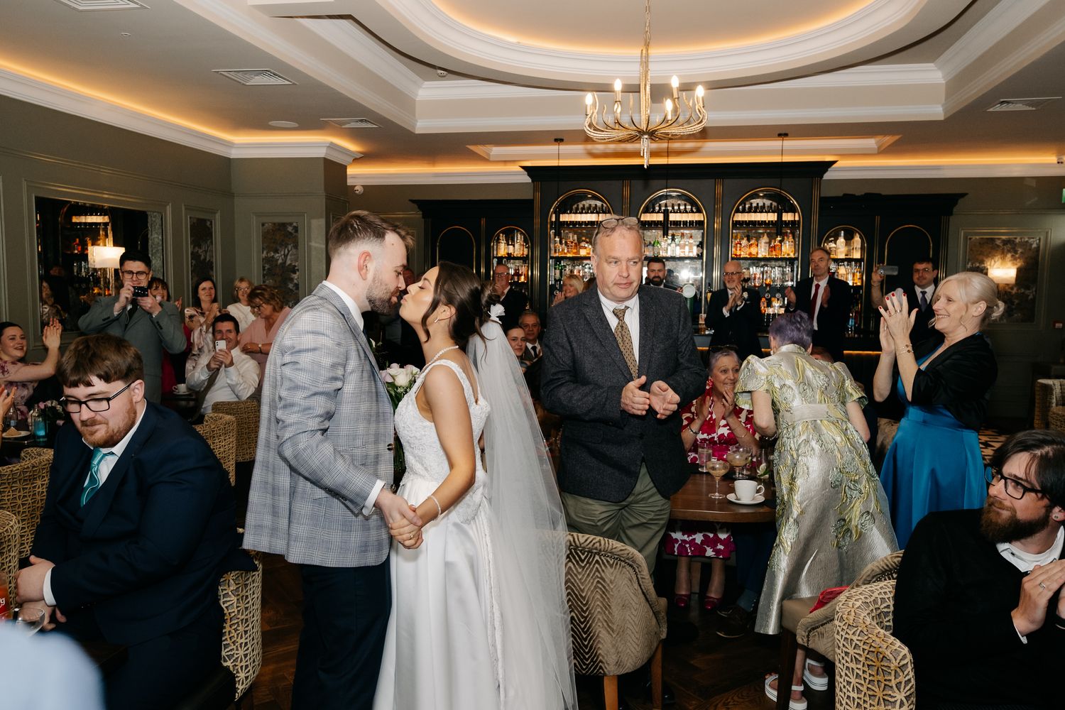 Bride and groom share a kiss during indoor wedding ceremony with guests celebrating in elegant venue.