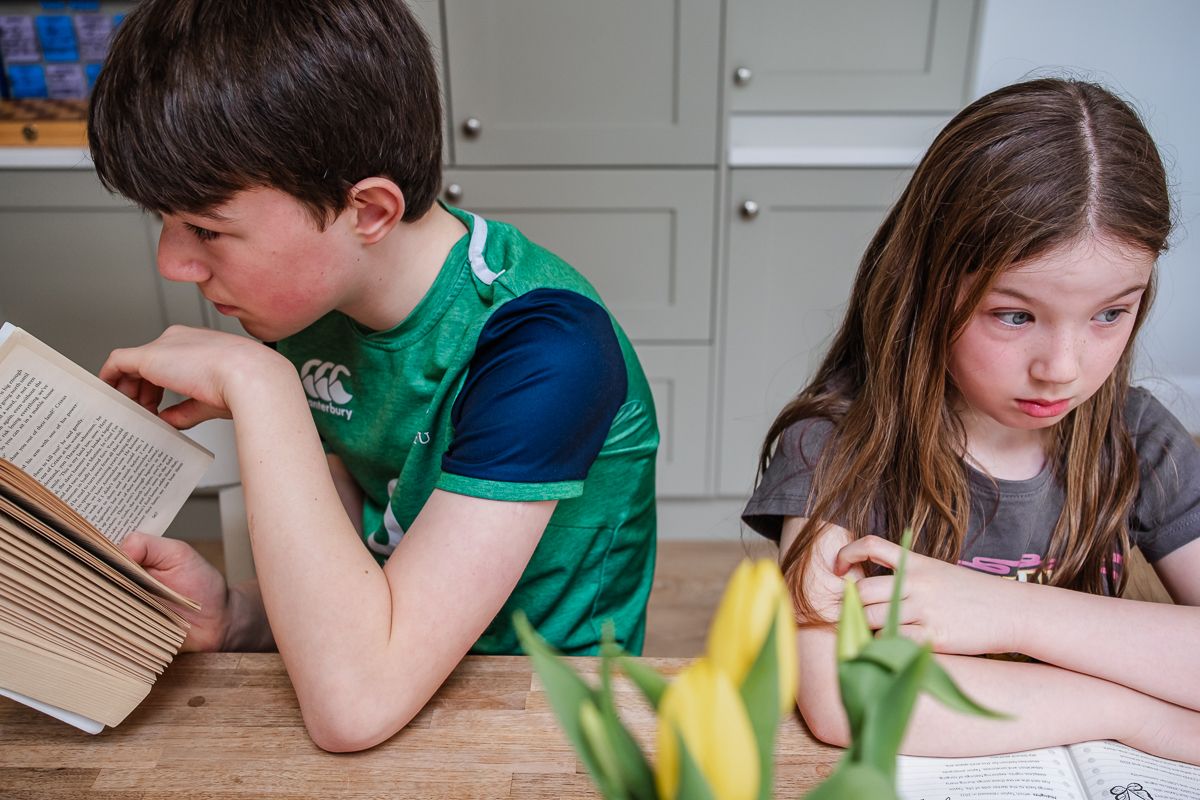 Two children sit at kitchen table, boy reading book and girl writing, with yellow tulips nearby.