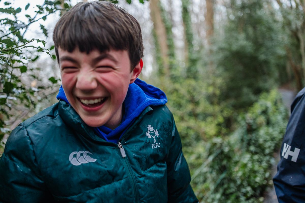 A laughing boy in a green Ireland rugby jacket enjoys an outdoor moment surrounded by lush greenery.