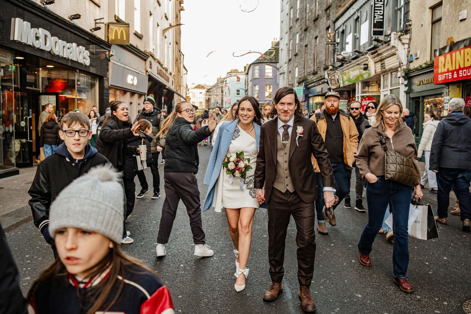 Bride and groom walk through busy city street holding bouquet, surrounded by cheering crowd near McDonald's.