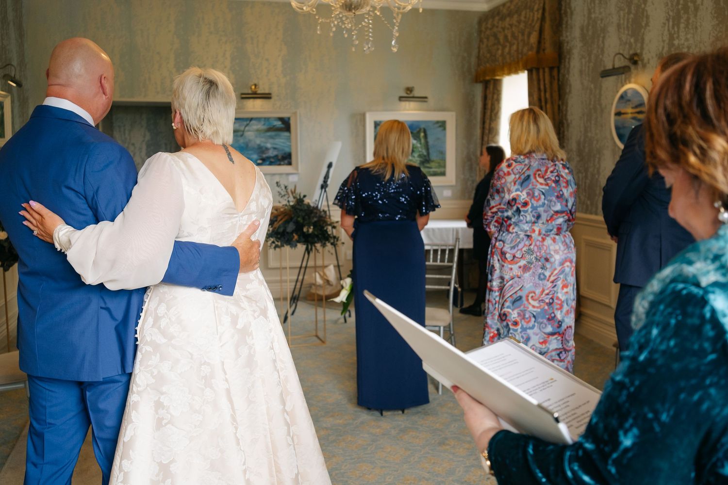 Bride and groom share a tender moment during intimate indoor wedding ceremony with guests and officiant present.